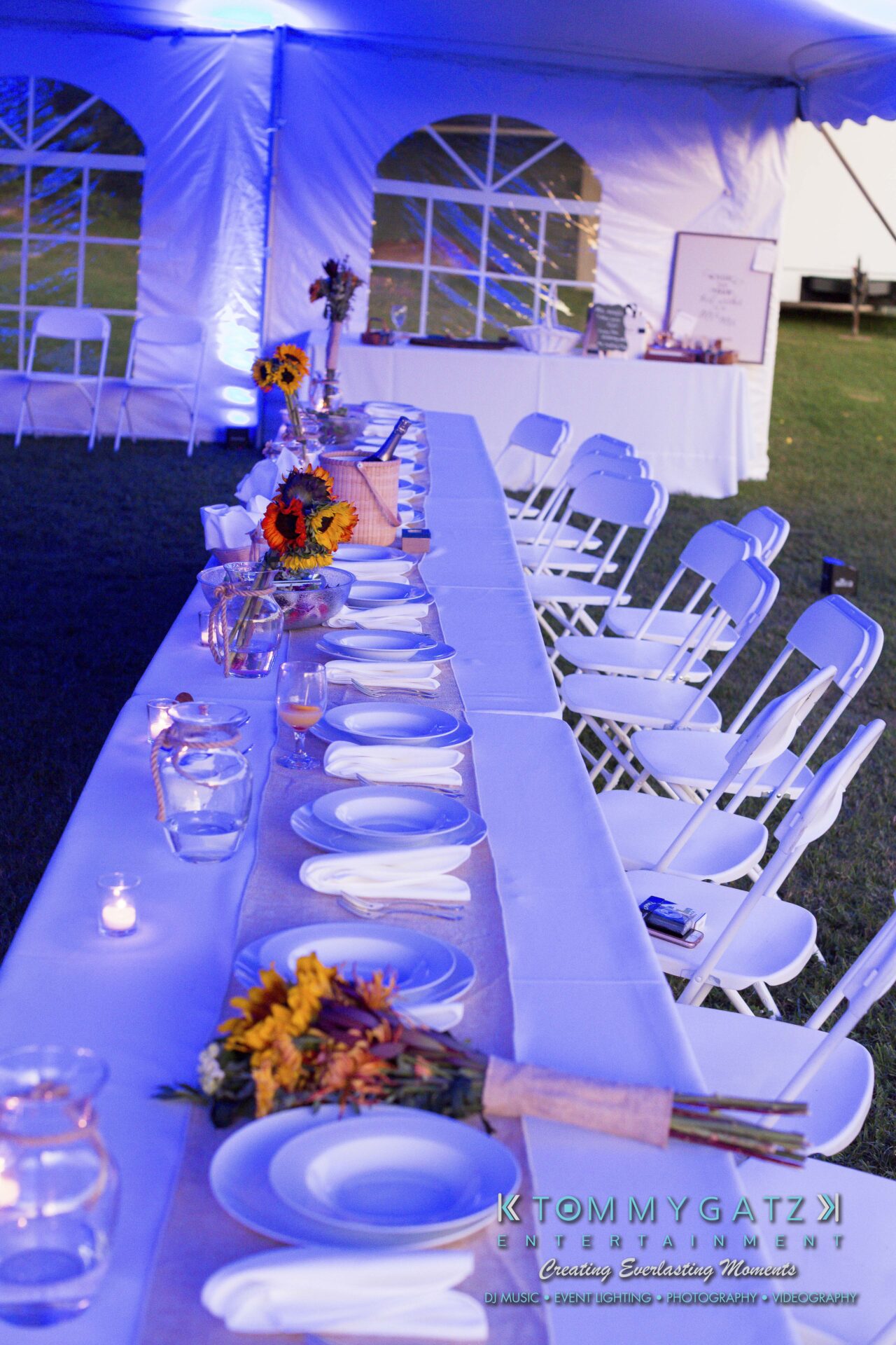 Ann-Marie-Alex-92317-RECEPTION-Low-Res-WM-13 - TOMMY GATZ ENTERTAINMENT A long, elegantly set table for an event with white chairs and glowing candles under a tent. Plates and napkins are arranged neatly, with floral centerpieces and ambient blue lighting.