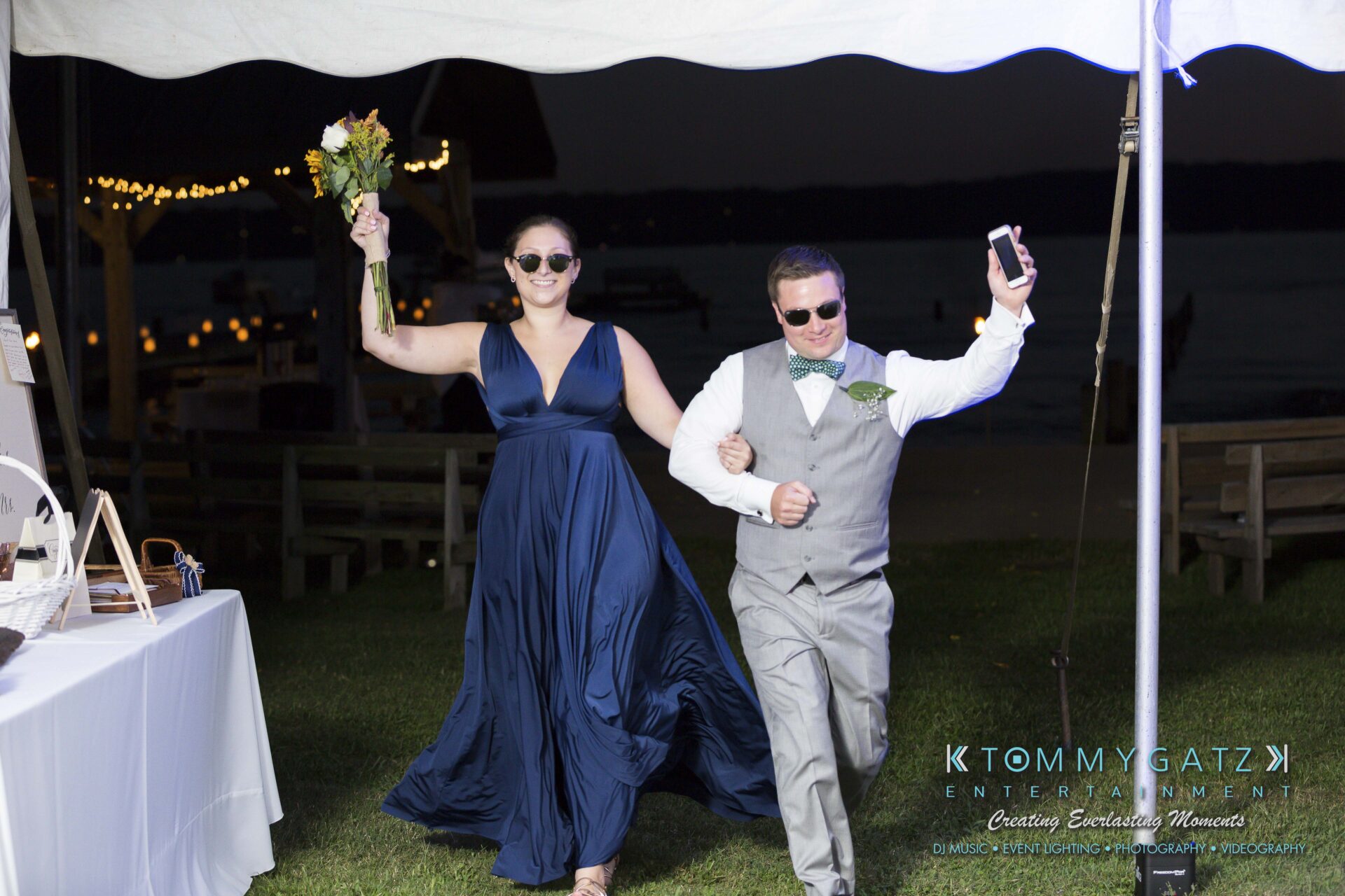 Ann-Marie-Alex-92317-RECEPTION-Low-Res-WM-16 - TOMMY GATZ ENTERTAINMENT A smiling man and woman, wearing sunglasses, walk arm in arm under a tent. The woman holds a bouquet, and the man holds a phone. They are dressed in formal attire.