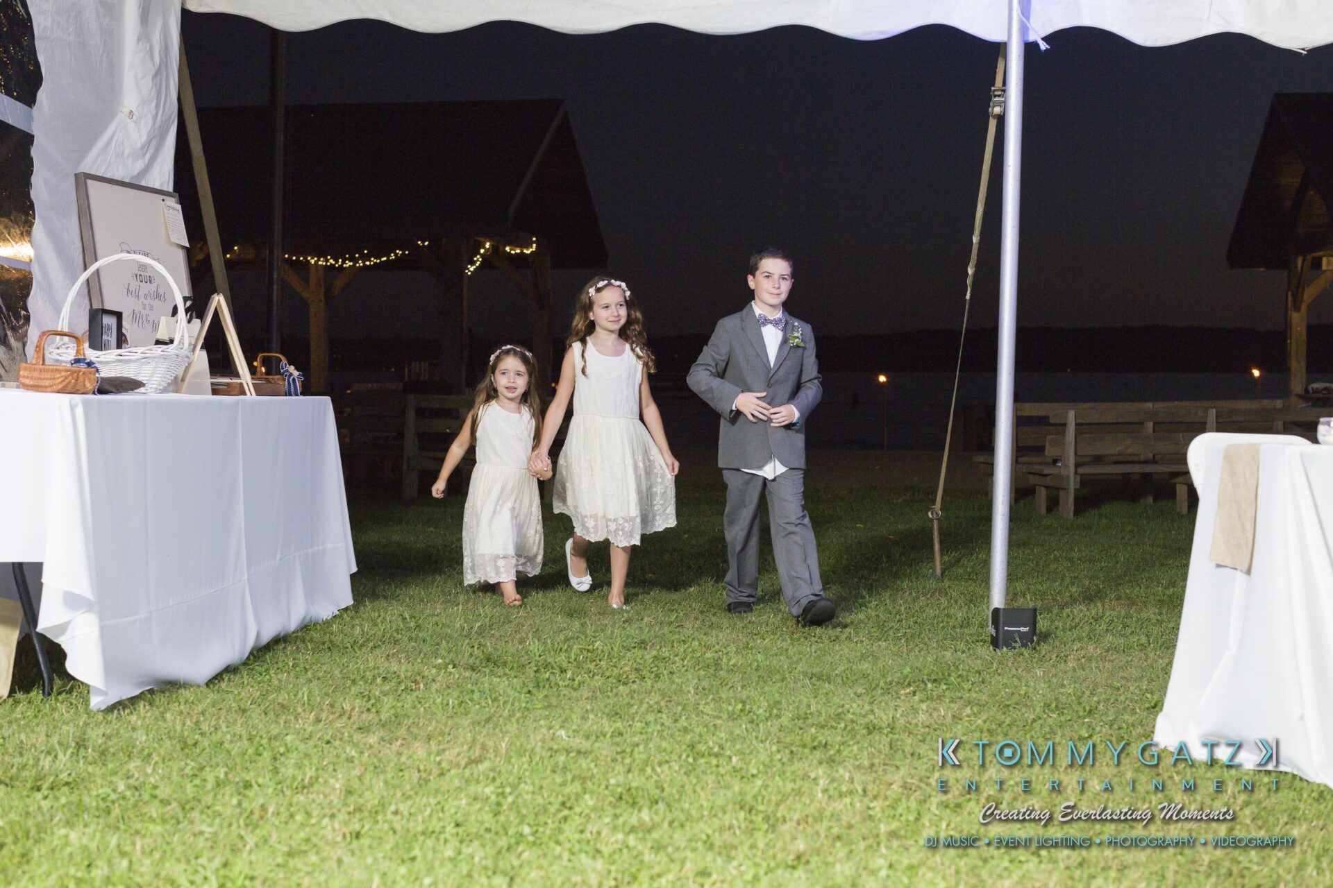 Ann-Marie-Alex-92317-RECEPTION-Low-Res-WM-21 - TOMMY GATZ ENTERTAINMENT Three children, two girls in white dresses and a boy in a suit, walk on grass under a tent at an outdoor evening event.