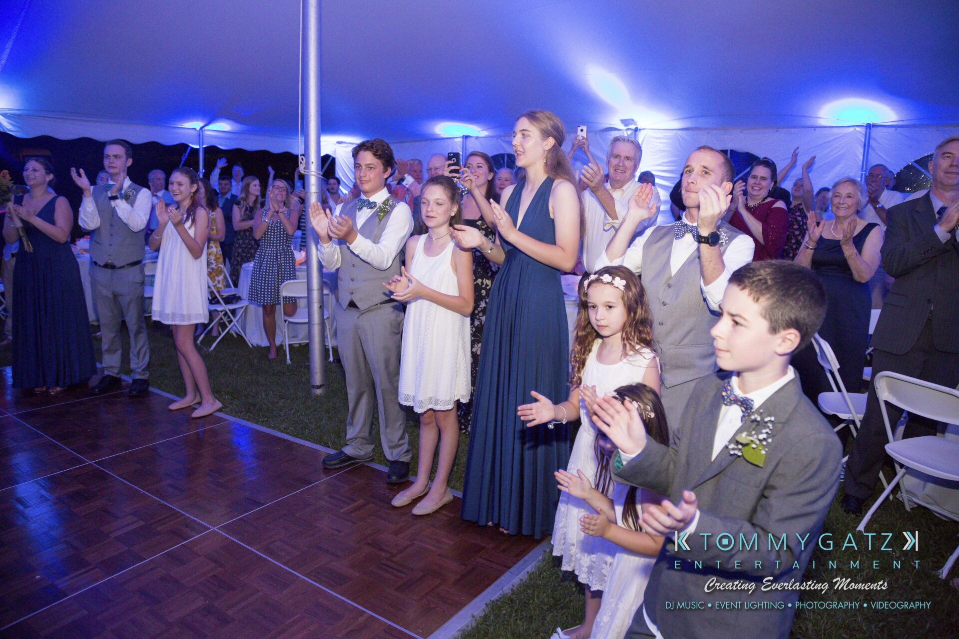 Ann-Marie-Alex-92317-RECEPTION-Low-Res-WM-22 - TOMMY GATZ ENTERTAINMENT A group of people, dressed formally, stands clapping under a tent with blue lighting.