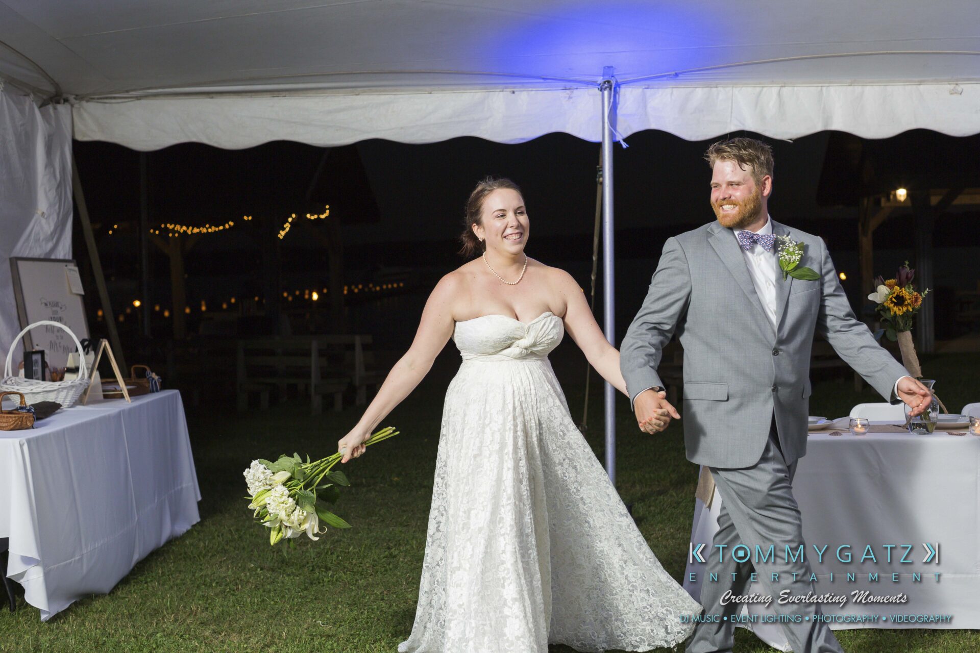 Ann-Marie-Alex-92317-RECEPTION-Low-Res-WM-26 - TOMMY GATZ ENTERTAINMENT A bride in a white gown and a groom in a gray suit enter a wedding reception tent, smiling and holding hands.