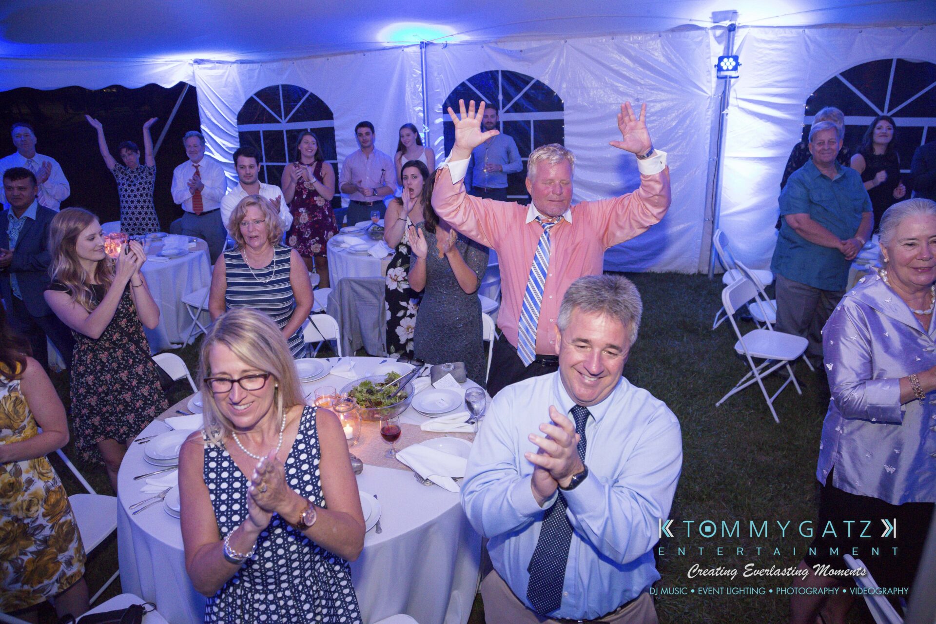 Ann-Marie-Alex-92317-RECEPTION-Low-Res-WM-28 - TOMMY GATZ ENTERTAINMENT A group of people at an indoor event, some seated at round tables and others standing, smiling and clapping. A banner for Tommy Gatz Entertainment is visible in the corner.