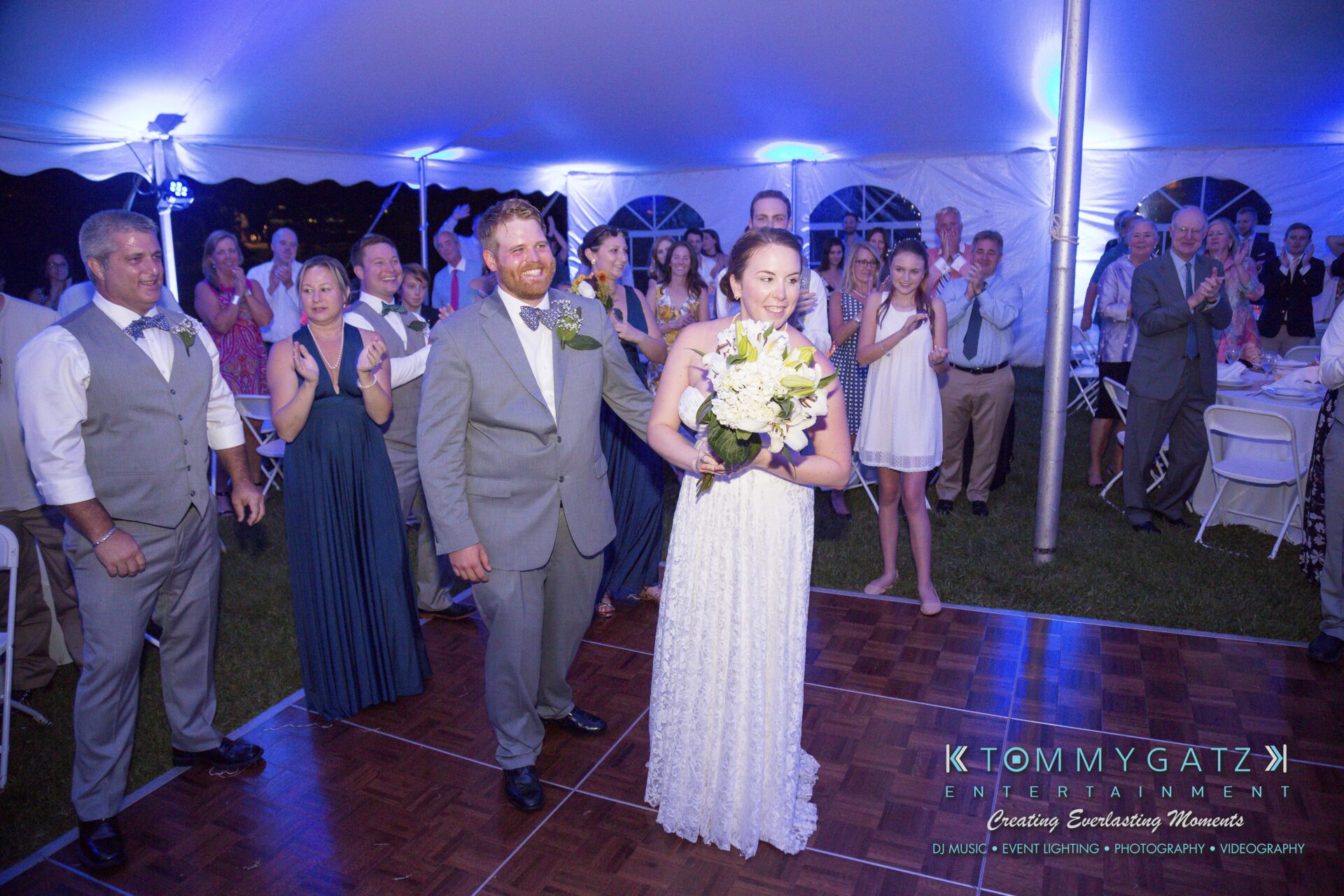 Ann-Marie-Alex-92317-RECEPTION-Low-Res-WM-34 - TOMMY GATZ ENTERTAINMENT A bride and groom stand on a dance floor under a white tent, surrounded by applauding guests. She holds a bouquet, and a sign for Tommy Gatz Entertainment is visible.
