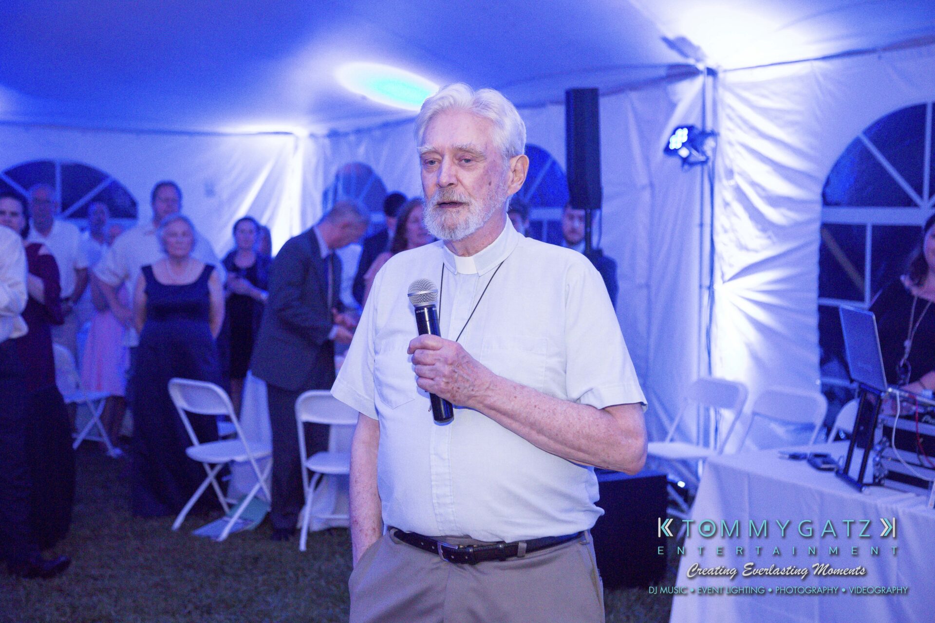 Ann-Marie-Alex-92317-RECEPTION-Low-Res-WM-35 - TOMMY GATZ ENTERTAINMENT An elderly man holding a microphone stands in a tent, addressing an audience seated on white chairs.