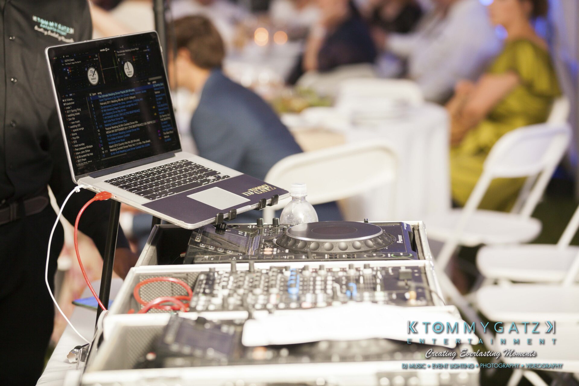 Ann-Marie-Alex-92317-RECEPTION-Low-Res-WM-36 - TOMMY GATZ ENTERTAINMENT DJ setup with a laptop and mixing console on a table at an event, with people seated in the background.