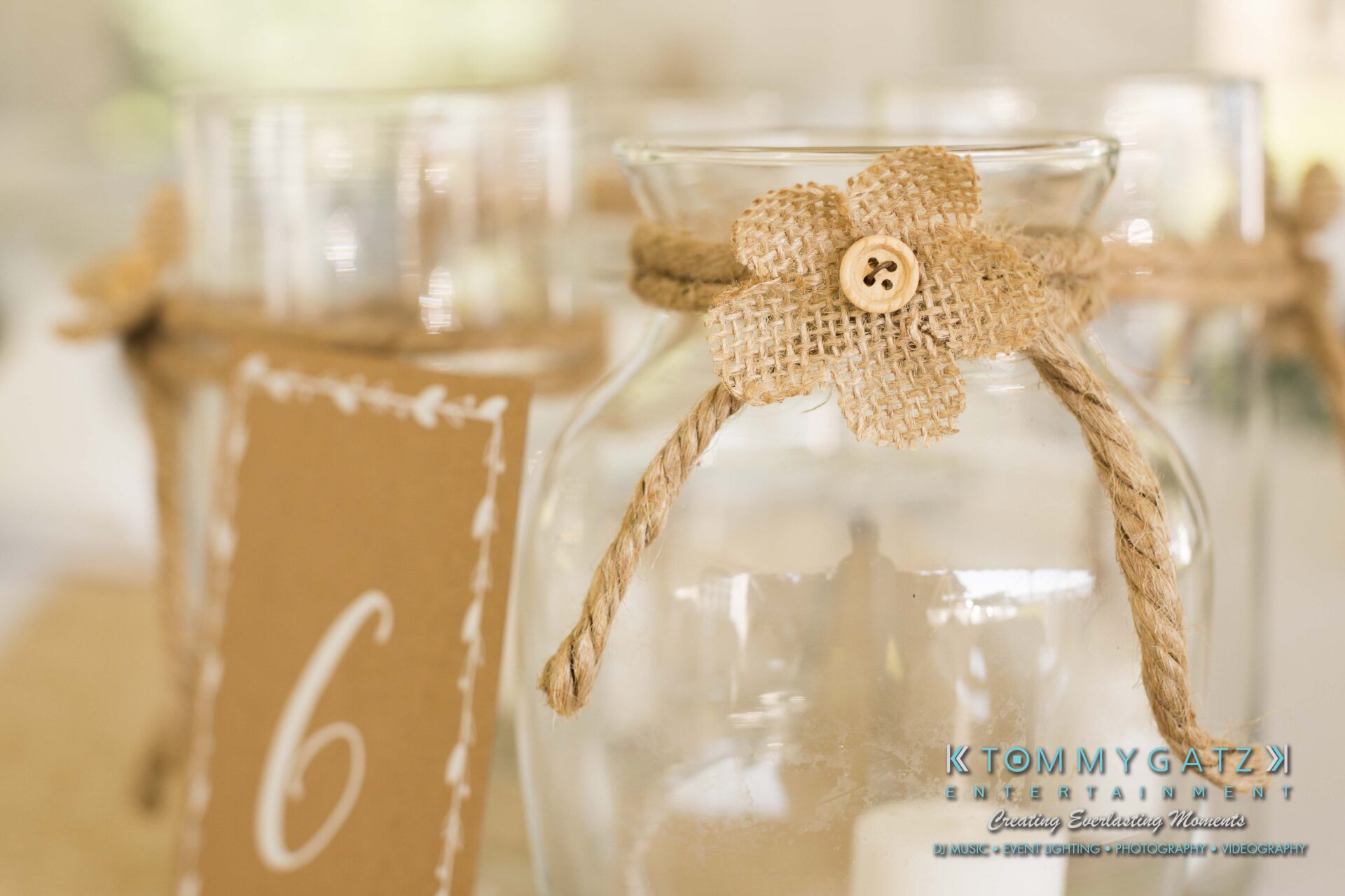 Ann-Marie-Alex-92317-RECEPTION-Low-Res-WM-4 - TOMMY GATZ ENTERTAINMENT Close-up of a decorative glass jar with a burlap flower and rope tie. A paper tag with the number six is visible in the background.