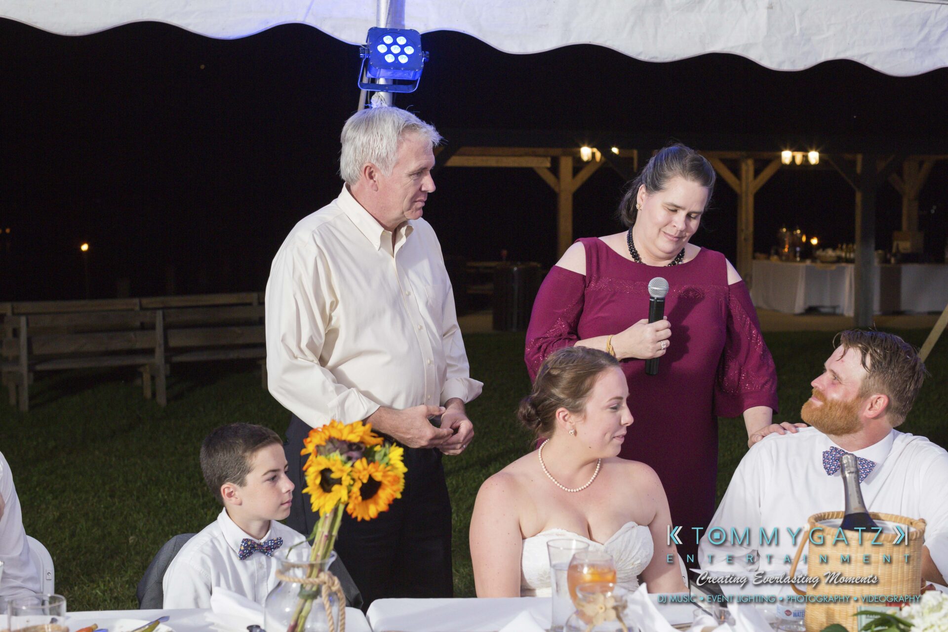 Ann-Marie-Alex-92317-RECEPTION-Low-Res-WM-42 - TOMMY GATZ ENTERTAINMENT A group of people at a formal event, with a woman holding a microphone, speaking to a seated couple. A young boy and an older man stand nearby, with a decorated table in the foreground.
