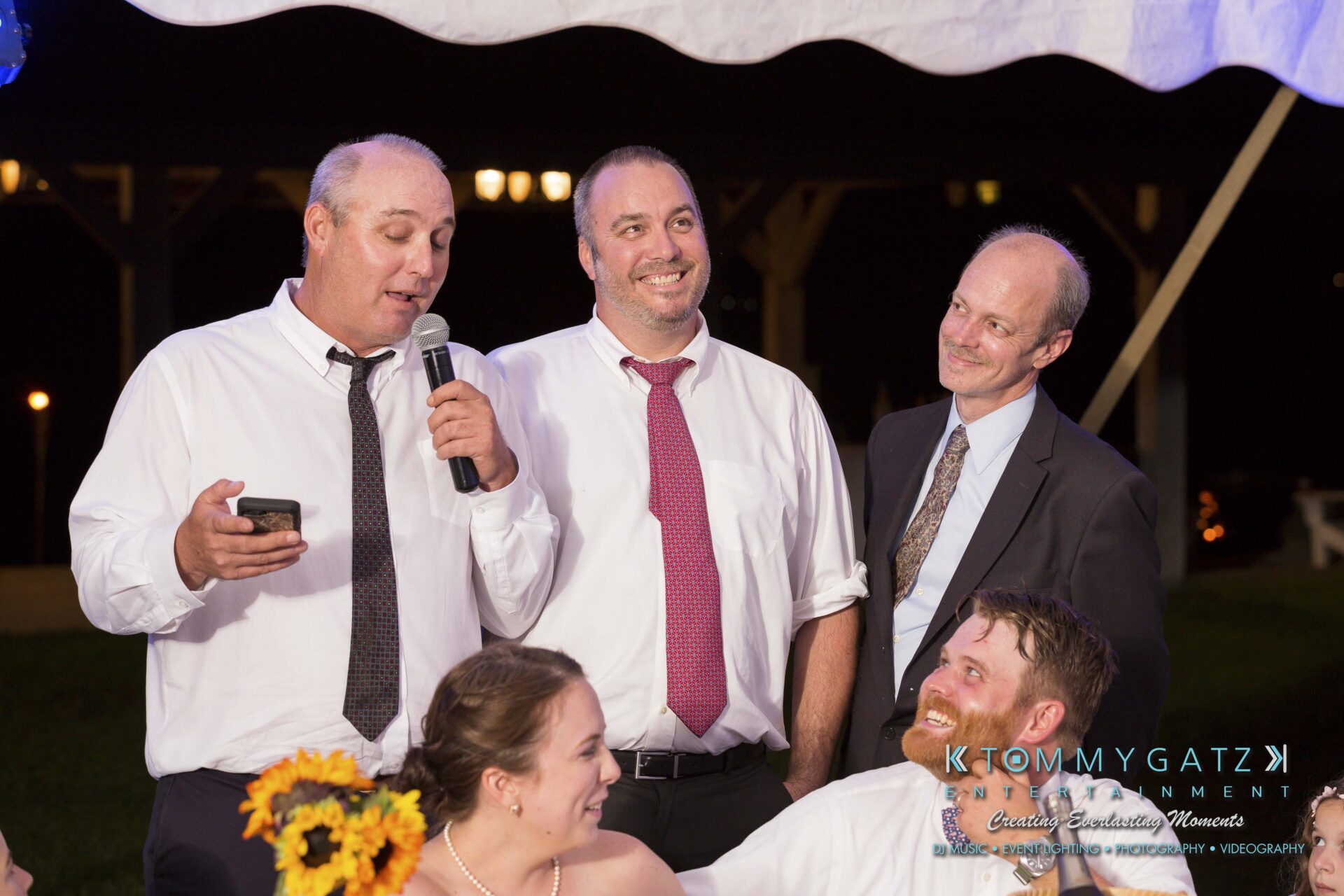 Ann-Marie-Alex-92317-RECEPTION-Low-Res-WM-44 - TOMMY GATZ ENTERTAINMENT Group of four men standing together under a tent; one holds a microphone while others smile. A seated couple at the bottom listens.