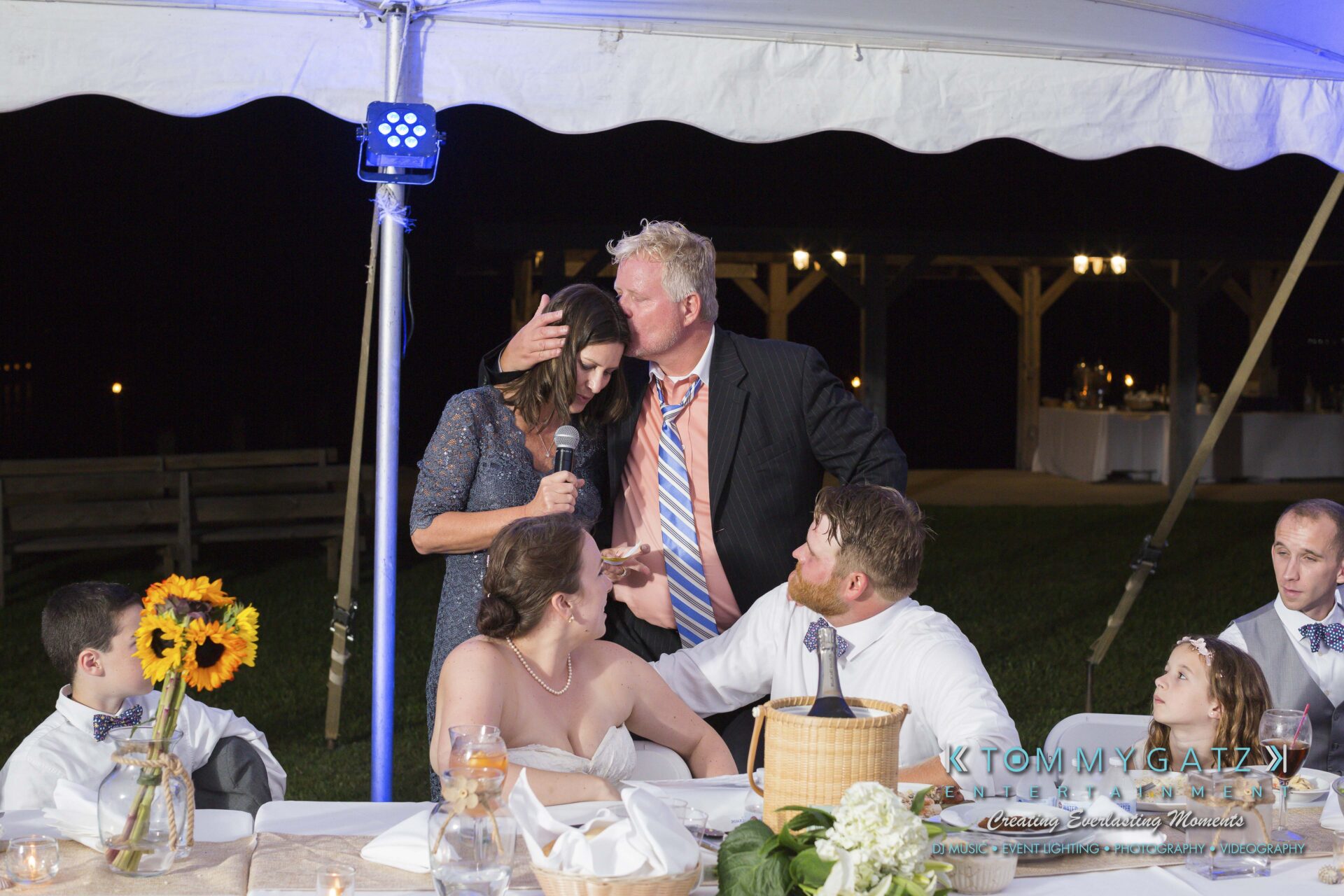 Ann-Marie-Alex-92317-RECEPTION-Low-Res-WM-50 - TOMMY GATZ ENTERTAINMENT A wedding reception under a tent with a group of people gathered around a table. A man in a suit gestures while others sit, some looking on with interest. A centerpiece of sunflowers is visible.