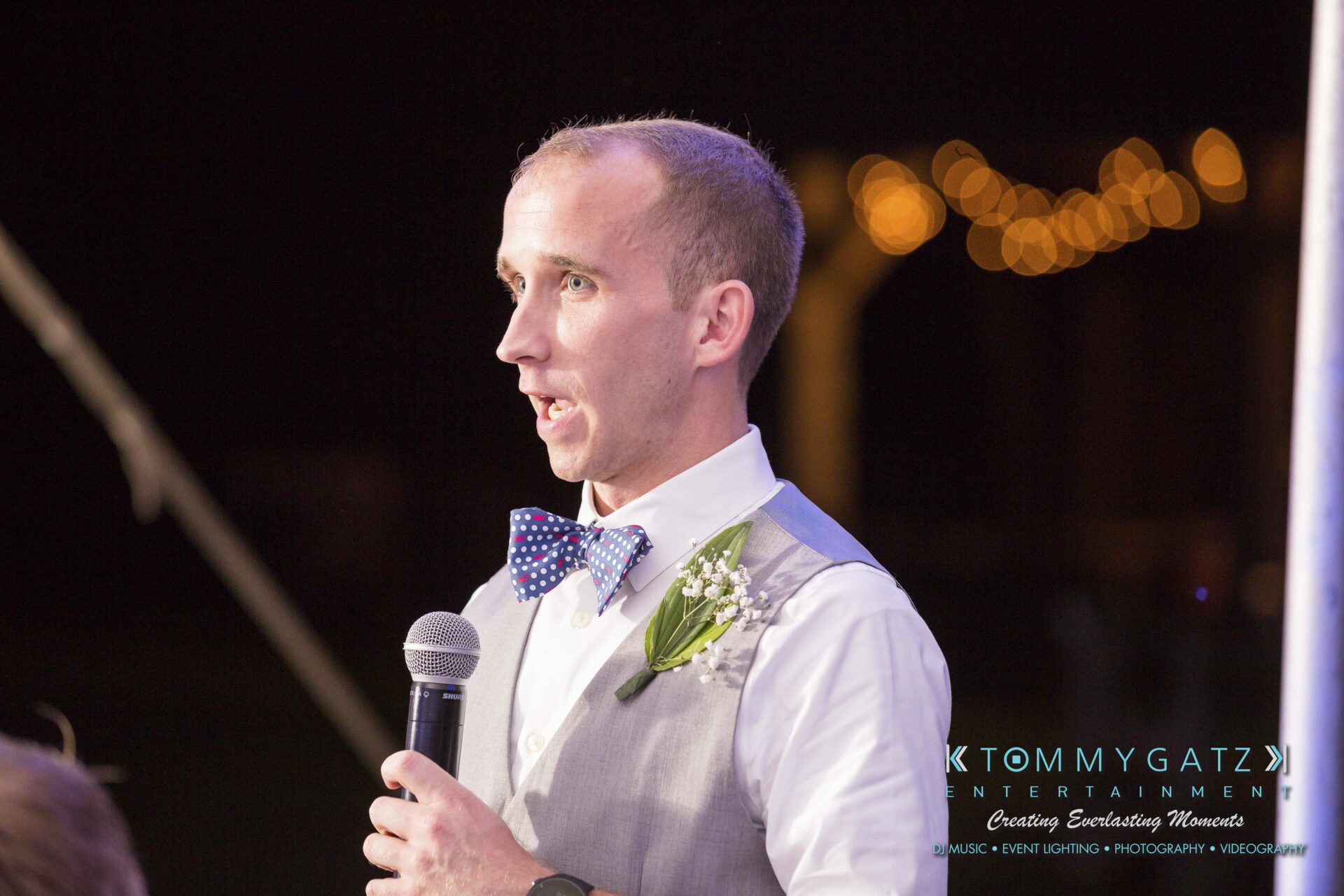 Ann-Marie-Alex-92317-RECEPTION-Low-Res-WM-51 - TOMMY GATZ ENTERTAINMENT A man in a bow tie and vest speaks into a microphone at an event with lights in the background.