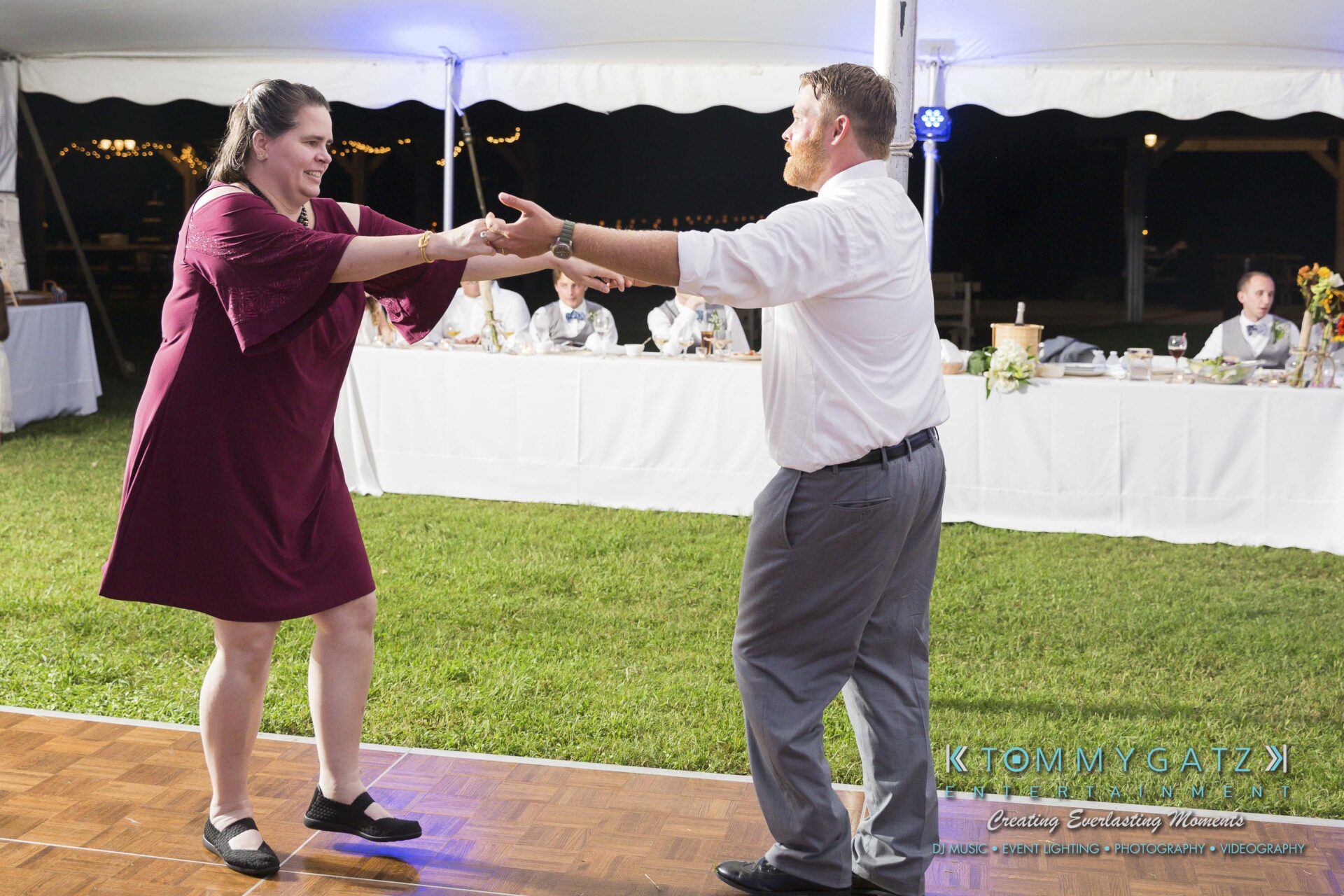 Ann-Marie-Alex-92317-RECEPTION-Low-Res-WM-66 - TOMMY GATZ ENTERTAINMENT A man and woman dance together on a wooden floor at an outdoor event, with people seated at a long table in the background.