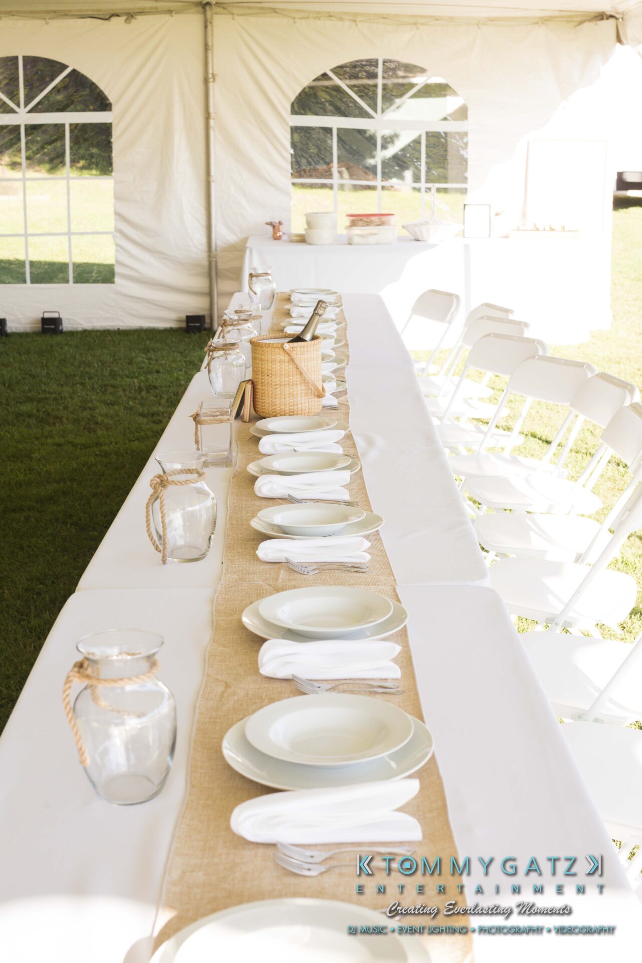 Ann-Marie-Alex-92317-RECEPTION-Low-Res-WM-7 - TOMMY GATZ ENTERTAINMENT A long table set for dining with white plates, napkins, and glass jars, under an outdoor tent with white chairs along both sides.