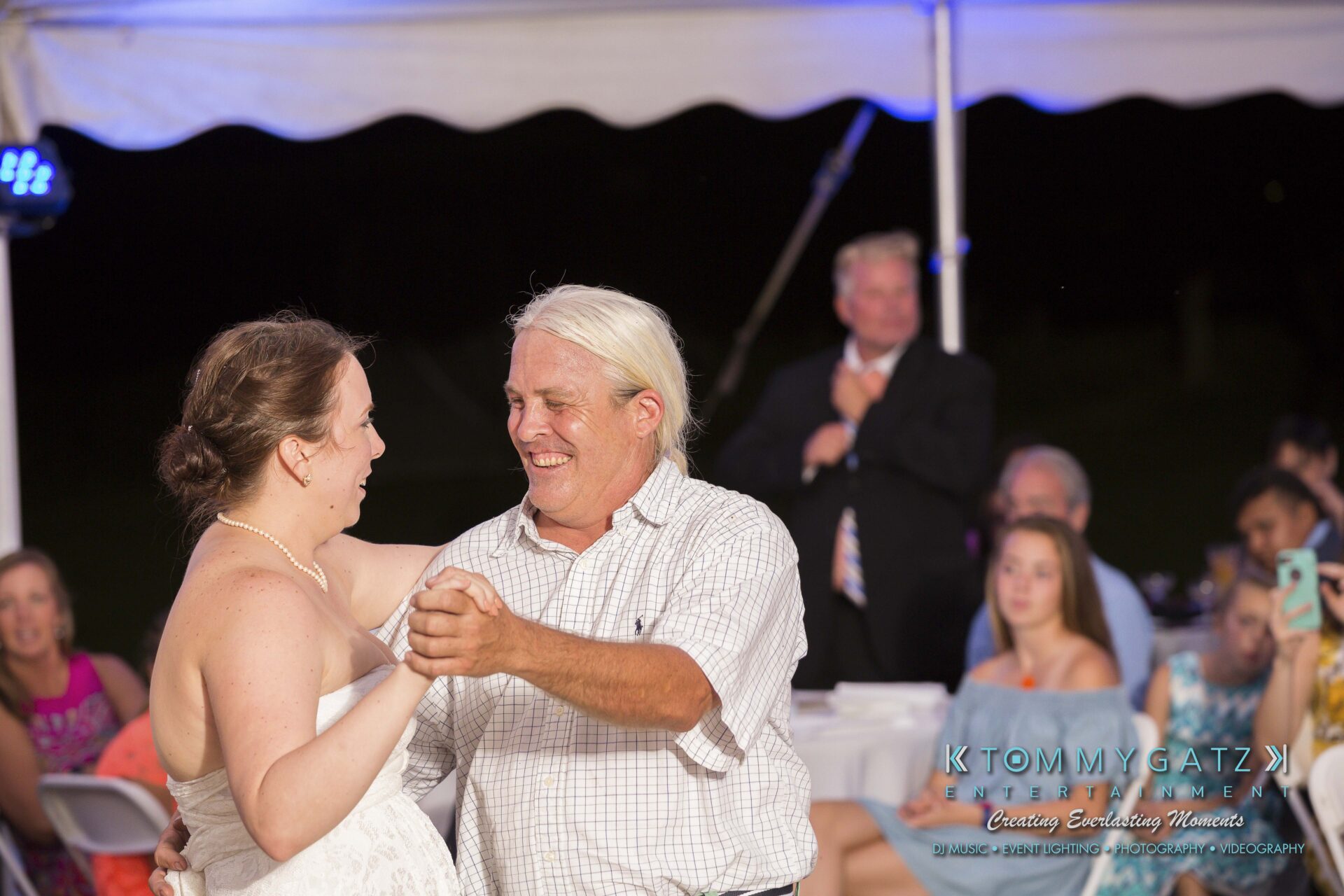 Ann-Marie-Alex-92317-RECEPTION-Low-Res-WM-74 - TOMMY GATZ ENTERTAINMENT A man and woman smiling while dancing together in an outdoor setting, with seated guests in the background.