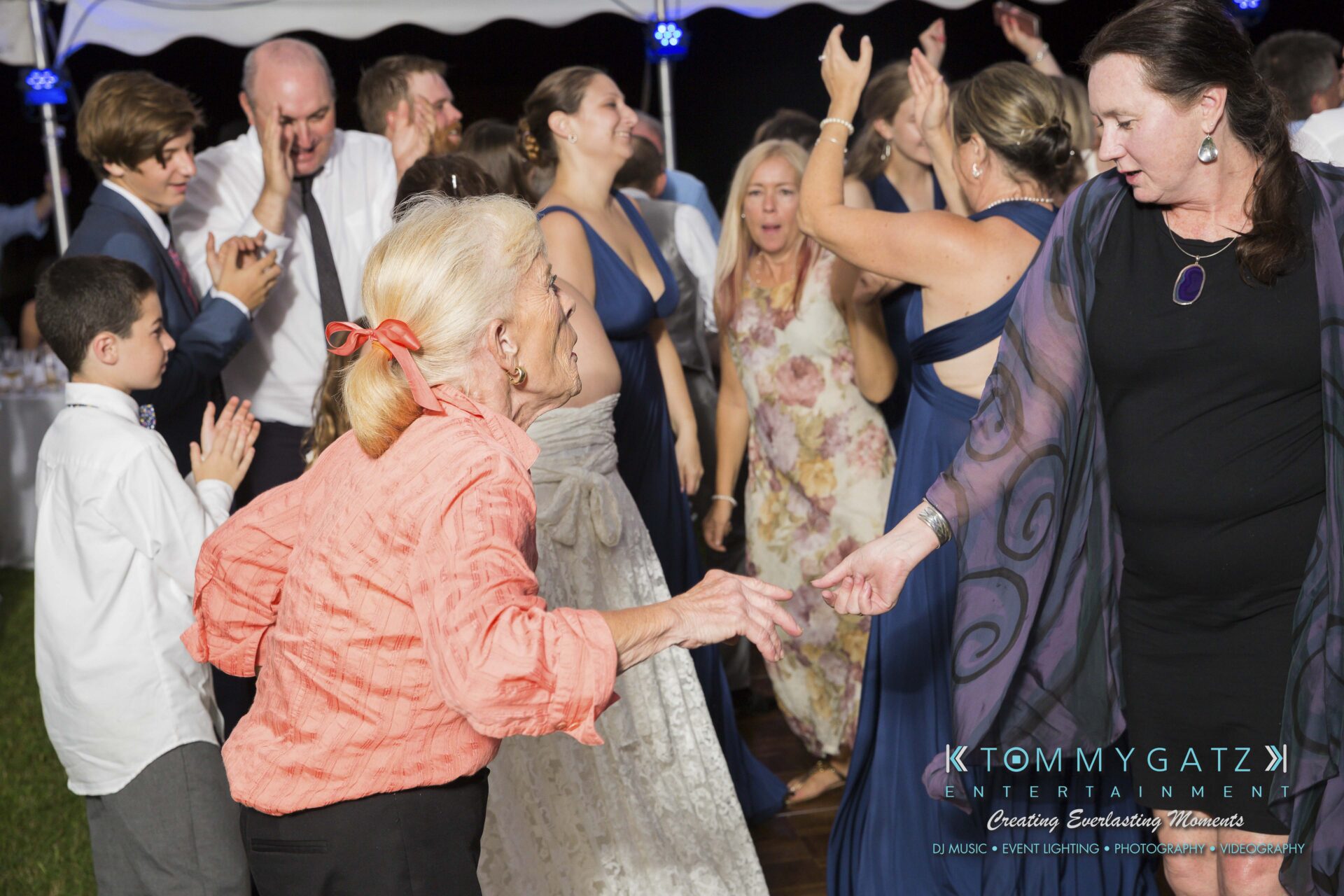 Ann-Marie-Alex-92317-RECEPTION-Low-Res-WM-85 - TOMMY GATZ ENTERTAINMENT People of various ages are dancing energetically at an outdoor event, with a focus on two women holding hands. The scene is lively and festive, and many guests are smiling.