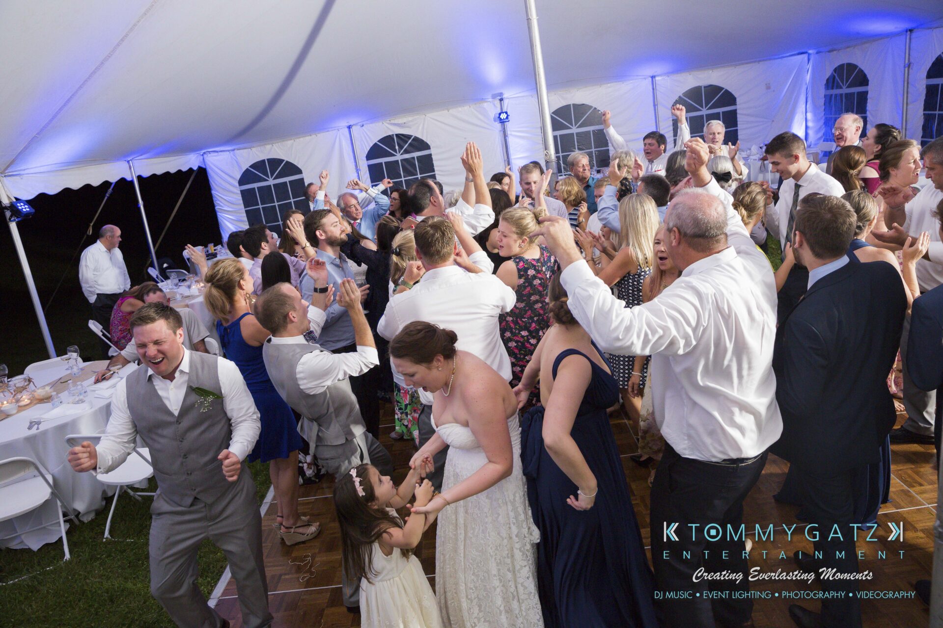 Ann-Marie-Alex-92317-RECEPTION-Low-Res-WM-87 - TOMMY GATZ ENTERTAINMENT Group of people dancing and celebrating under a white tent, including a bride in a wedding dress and flower girls.