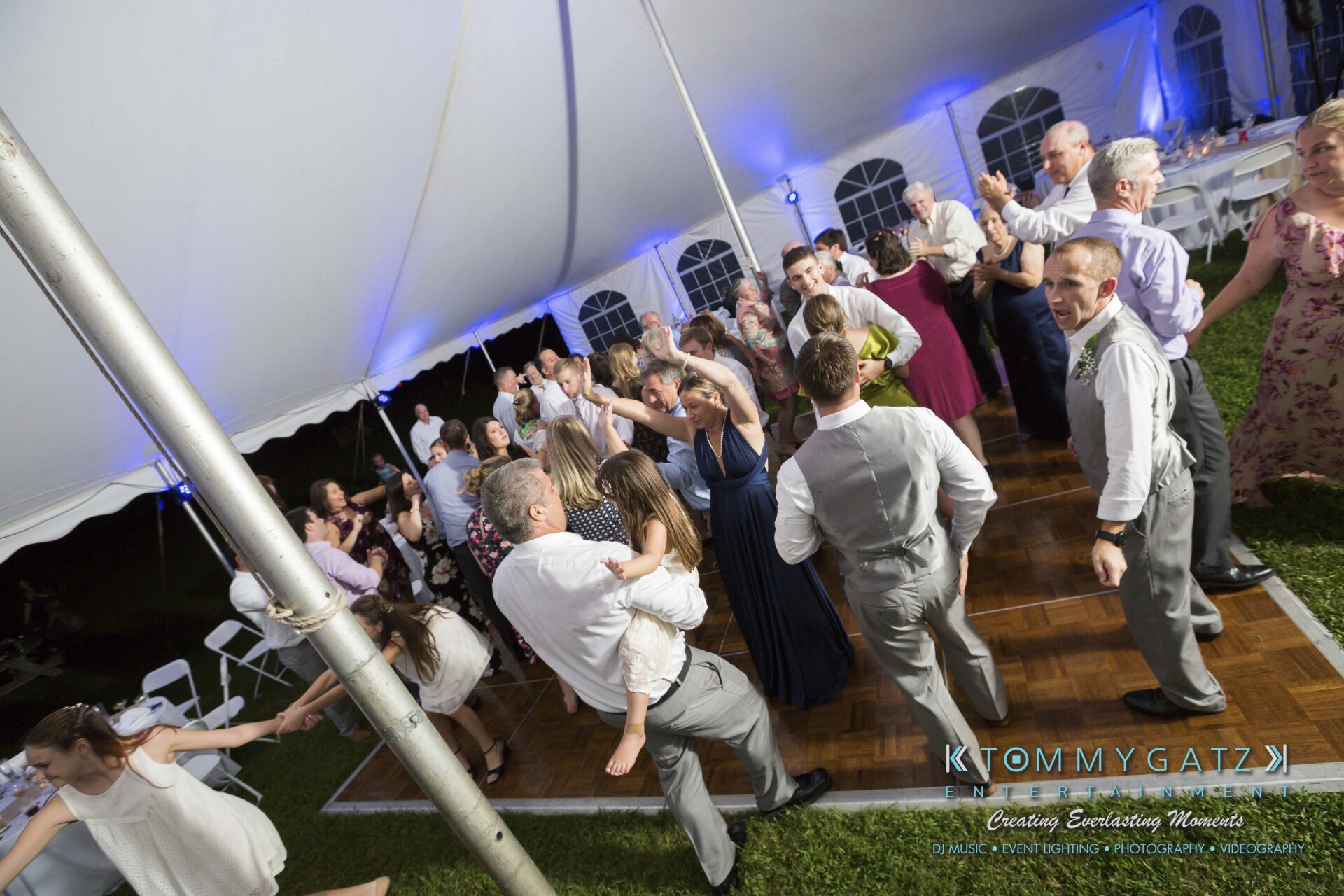 Ann-Marie-Alex-92317-RECEPTION-Low-Res-WM-93 - TOMMY GATZ ENTERTAINMENT A group of people dancing under a large white tent at an event. Some are seated, others are standing on a wooden dance floor. The atmosphere is lively and festive.