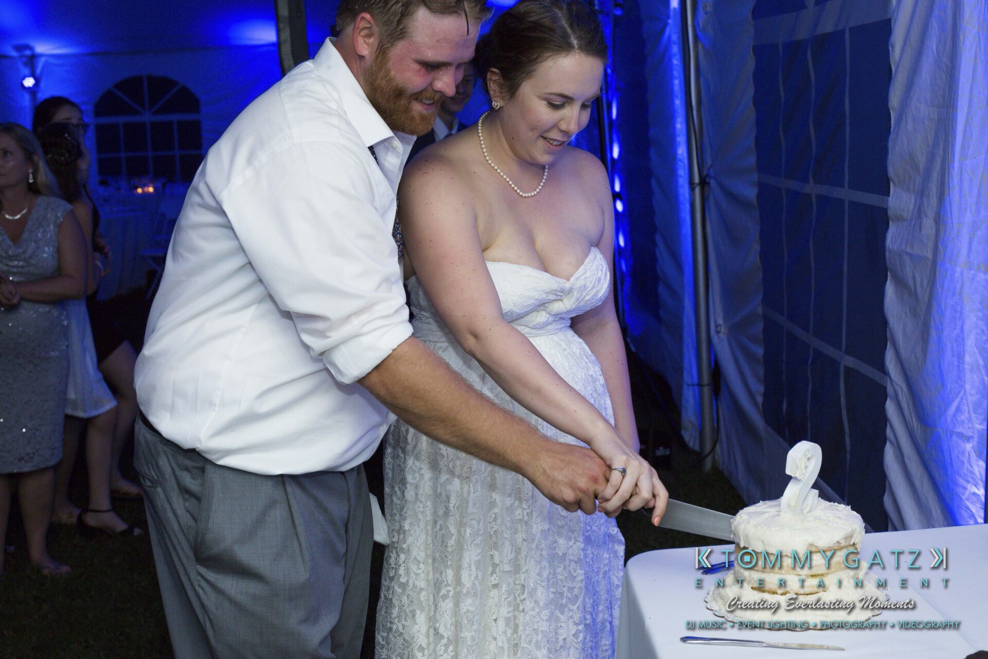 Ann-Marie-Alex-92317-RECEPTION-Low-Res-WM-96 - TOMMY GATZ ENTERTAINMENT A bride and groom cut a tiered wedding cake together under a blue-lit tent.