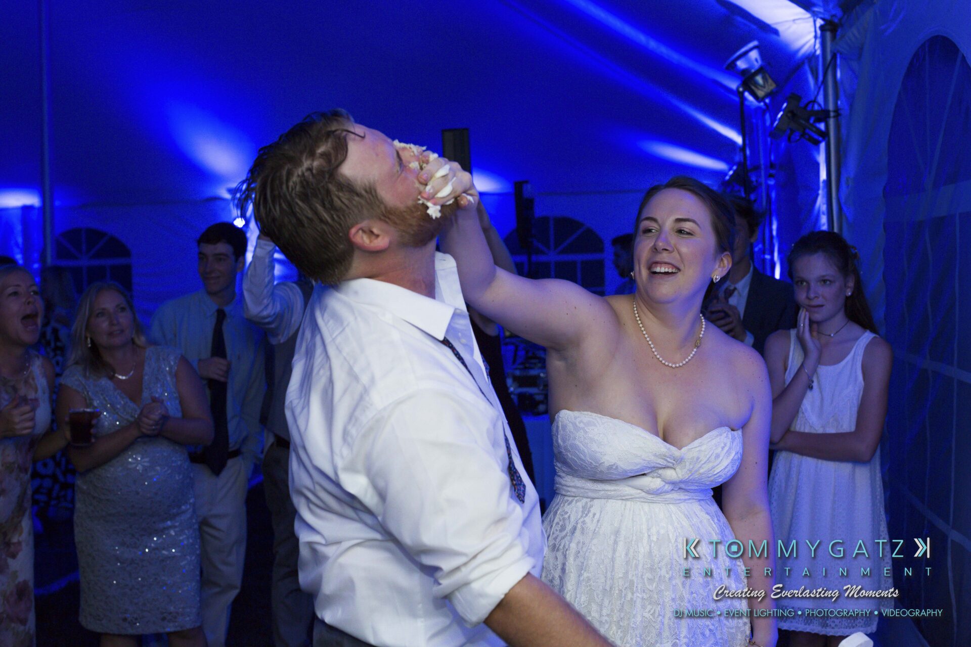 Ann-Marie-Alex-92317-RECEPTION-Low-Res-WM-98 - TOMMY GATZ ENTERTAINMENT A couple playfully smears cake on each other's faces at a wedding reception, surrounded by laughing guests in a blue-lit tent.