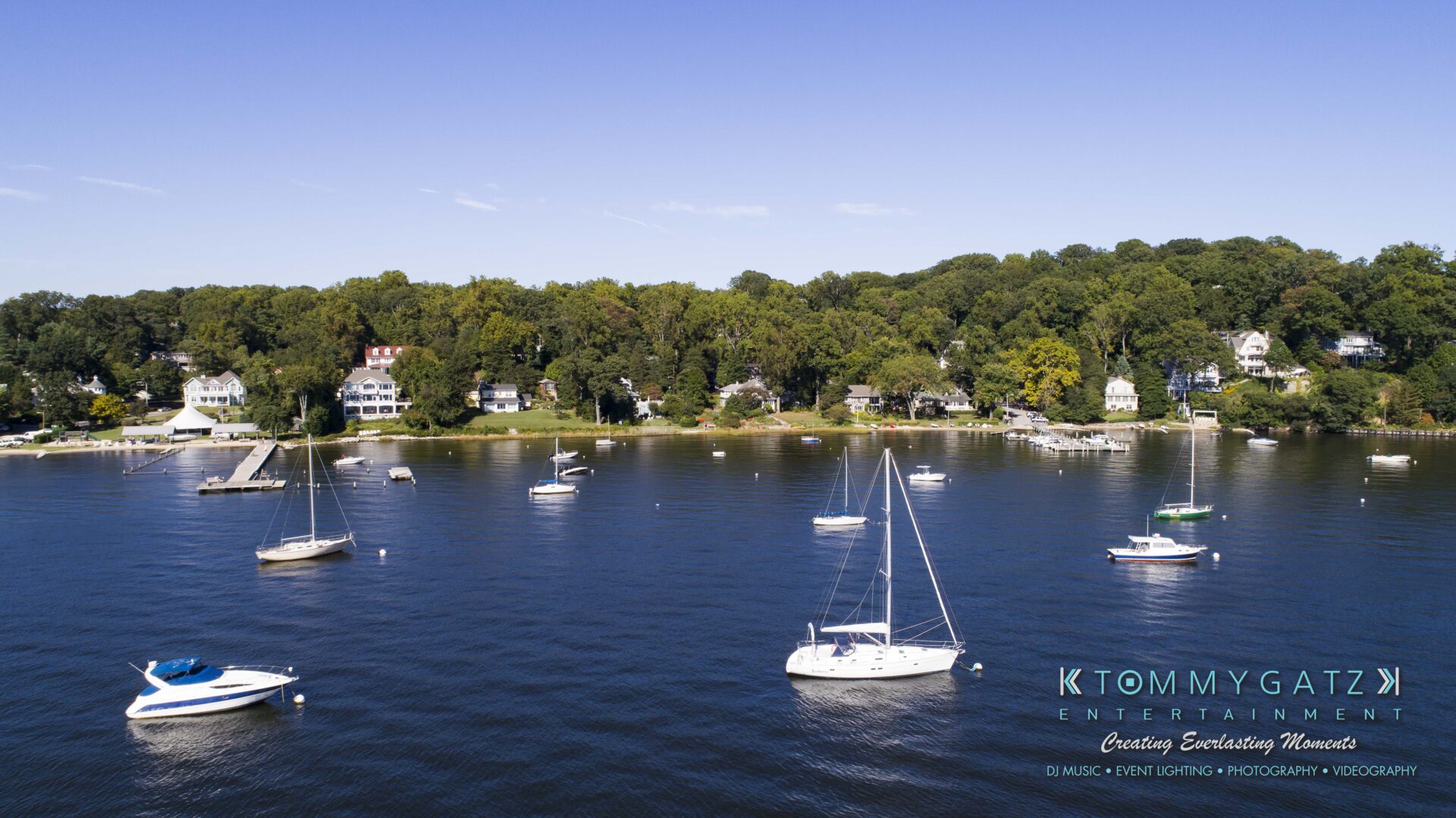 Ann-Marie-Alex-DRONE-Low-Res-WM-1 - TOMMY GATZ ENTERTAINMENT A serene coastal scene with several sailboats anchored in calm water, surrounded by lush green trees and waterfront houses.