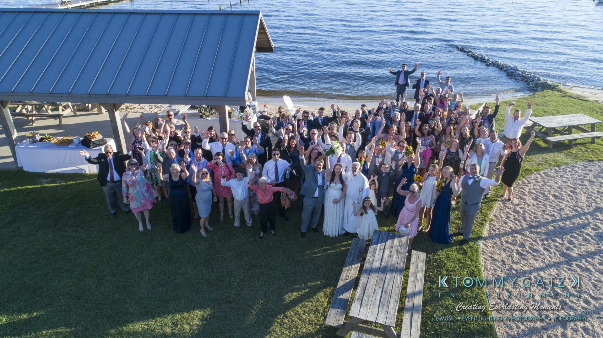 Aerial view of a large group gathered for a wedding celebration near a lakeside pavilion, with some waving and posing.