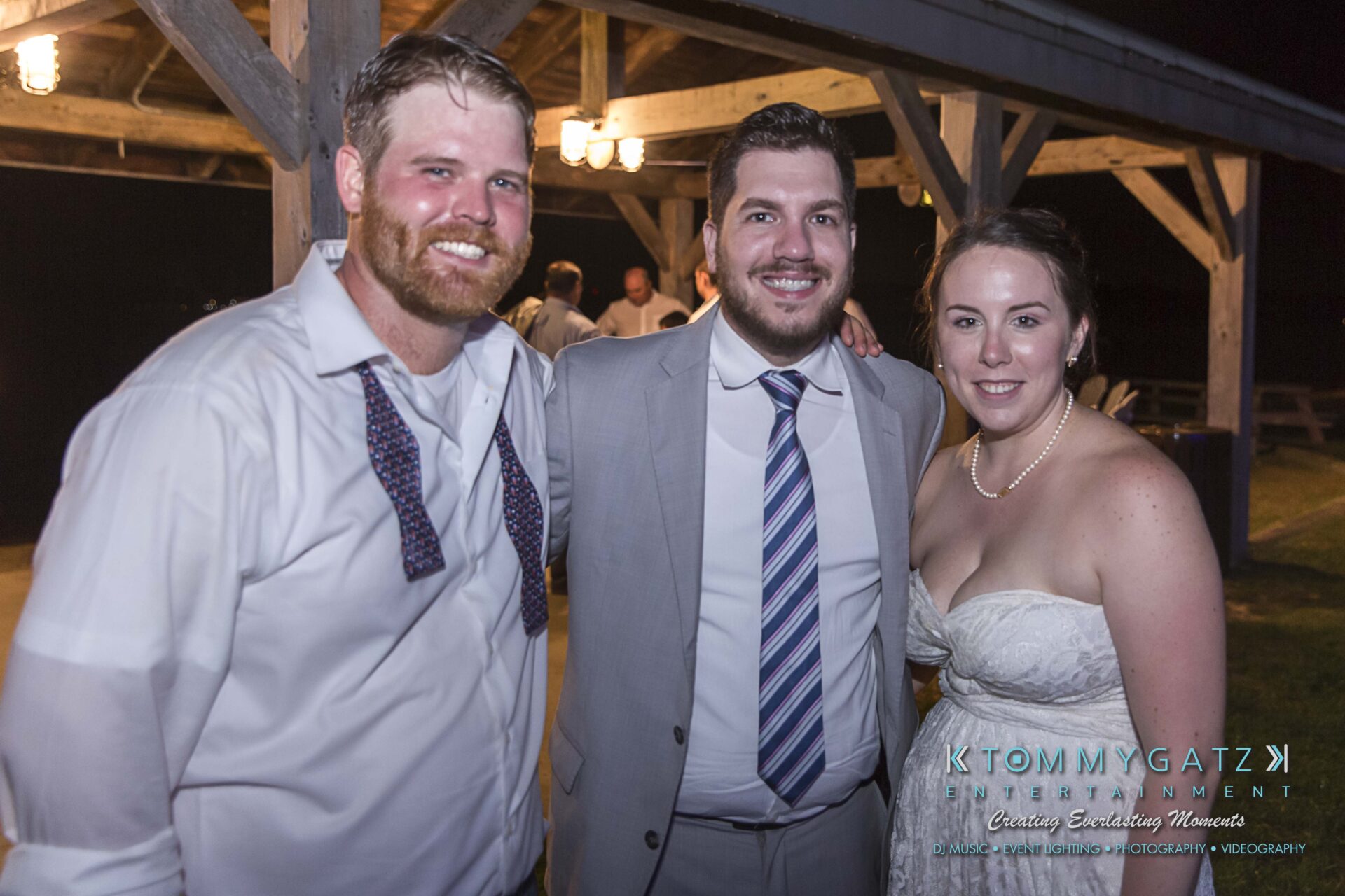 Ann-Marie-Alex-RECEPTION-Low-Res-WM-133 - TOMMY GATZ ENTERTAINMENT Three people posing together at an event. Two men are in dress shirts and ties, and a woman is in a white strapless dress. They are under a wooden pavilion with lights.