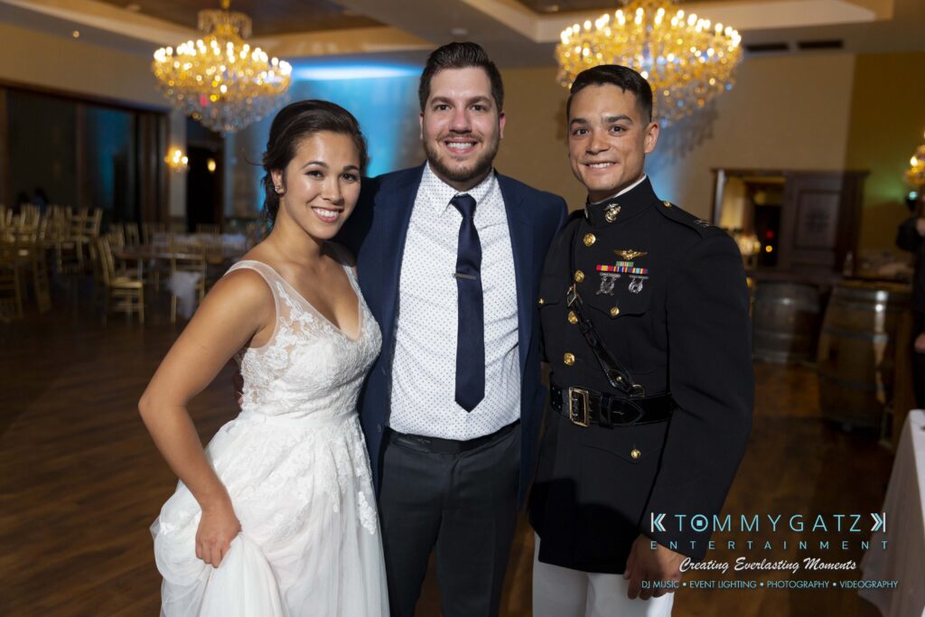 Annie-Casey-060918-CEREMONY-Low-Res-WM-424 - TOMMY GATZ ENTERTAINMENT Bride in white dress, two men in suits, one in military uniform, standing together in a banquet hall with chandeliers.