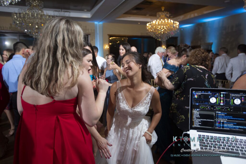 Annie-Casey-060918-RECEPTION-Low-Res-WM-1029 - TOMMY GATZ ENTERTAINMENT A bride in a white dress dances with guests at a wedding reception. The room is crowded, and there is a DJ setup with a laptop visible in the foreground.
