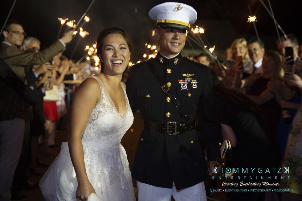 Annie-Casey-060918-RECEPTION-Low-Res-WM-1080 - TOMMY GATZ ENTERTAINMENT A bride and a groom in a military uniform smile surrounded by guests holding sparklers at an evening celebration.
