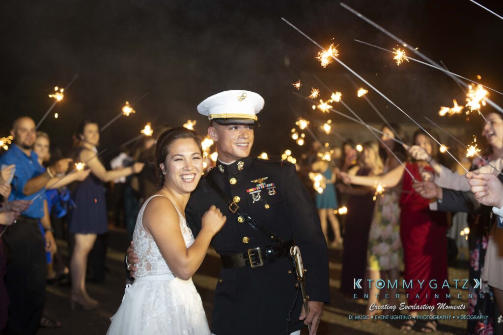 Annie-Casey-060918-RECEPTION-Low-Res-WM-1088 - TOMMY GATZ ENTERTAINMENT A couple, with the man in a dress uniform and the woman in a white dress, smiles surrounded by people holding sparklers.