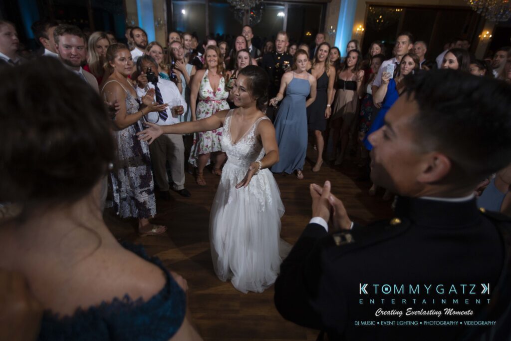 Annie-Casey-060918-RECEPTION-Low-Res-WM-990 - TOMMY GATZ ENTERTAINMENT Bride in a white dress dancing in the center, surrounded by a crowd of guests in a dimly lit ballroom.