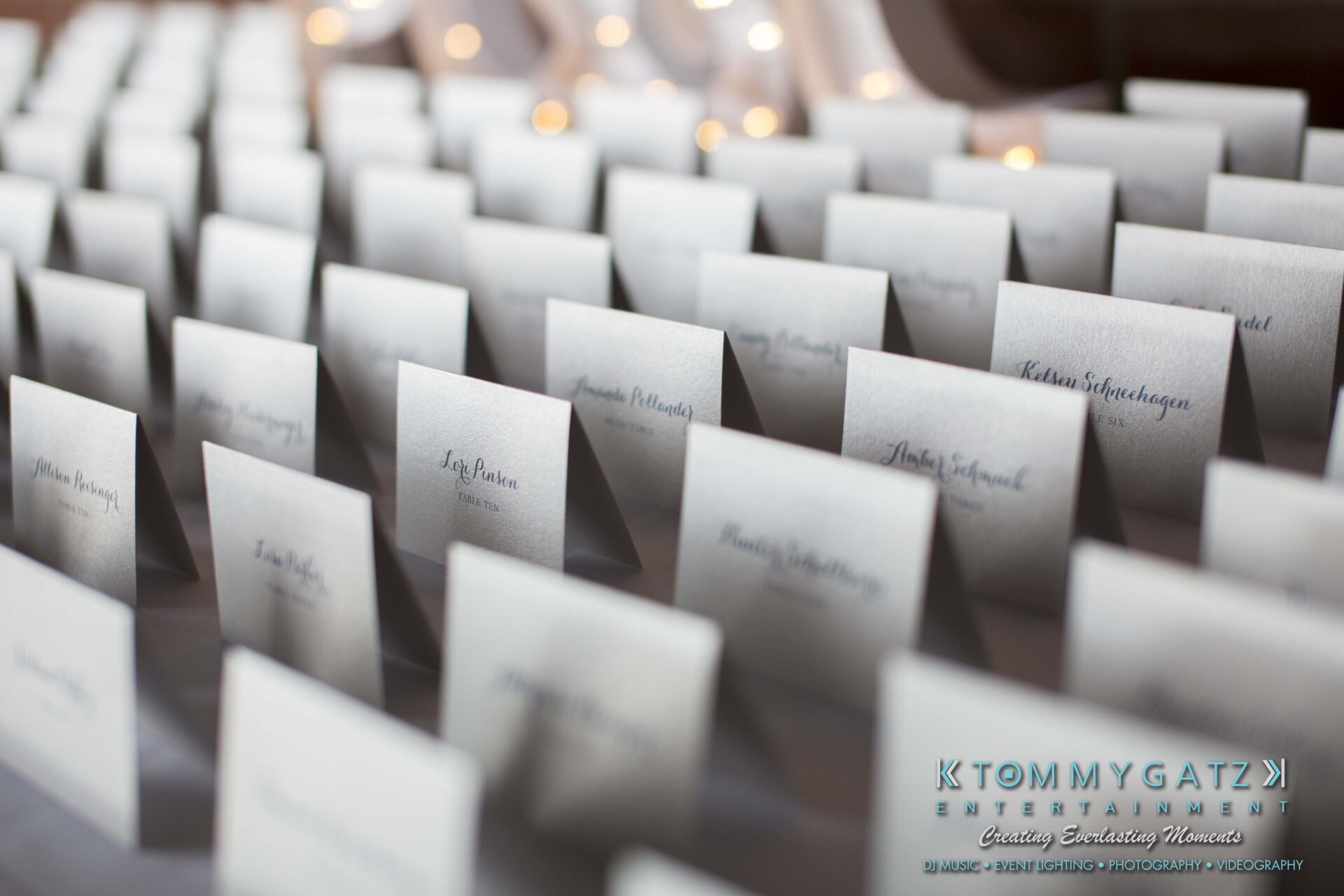 Carly-Patrick-FINAL-Blog-Watermark-1 - TOMMY GATZ ENTERTAINMENT Rows of white name cards arranged in a grid, each with a printed name, on a table decorated with small lights above.
