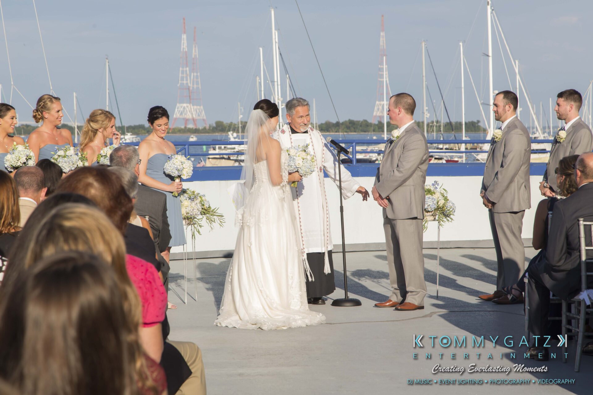 Carly-Patrick-FINAL-Blog-Watermark-11 - TOMMY GATZ ENTERTAINMENT A couple exchanges vows in an outdoor wedding ceremony. Bridesmaids and groomsmen stand on either side, with guests seated in the foreground. A body of water and towers are in the background.