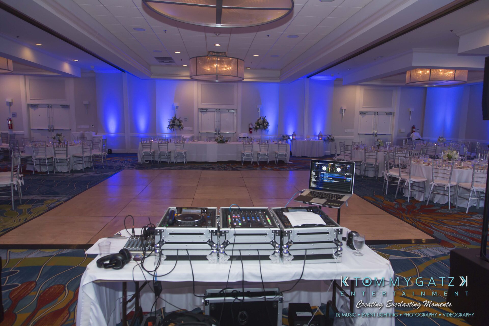 Carly-Patrick-FINAL-Blog-Watermark-12 - TOMMY GATZ ENTERTAINMENT DJ setup in a large banquet hall with blue lighting, empty round tables, and a wooden dance floor.