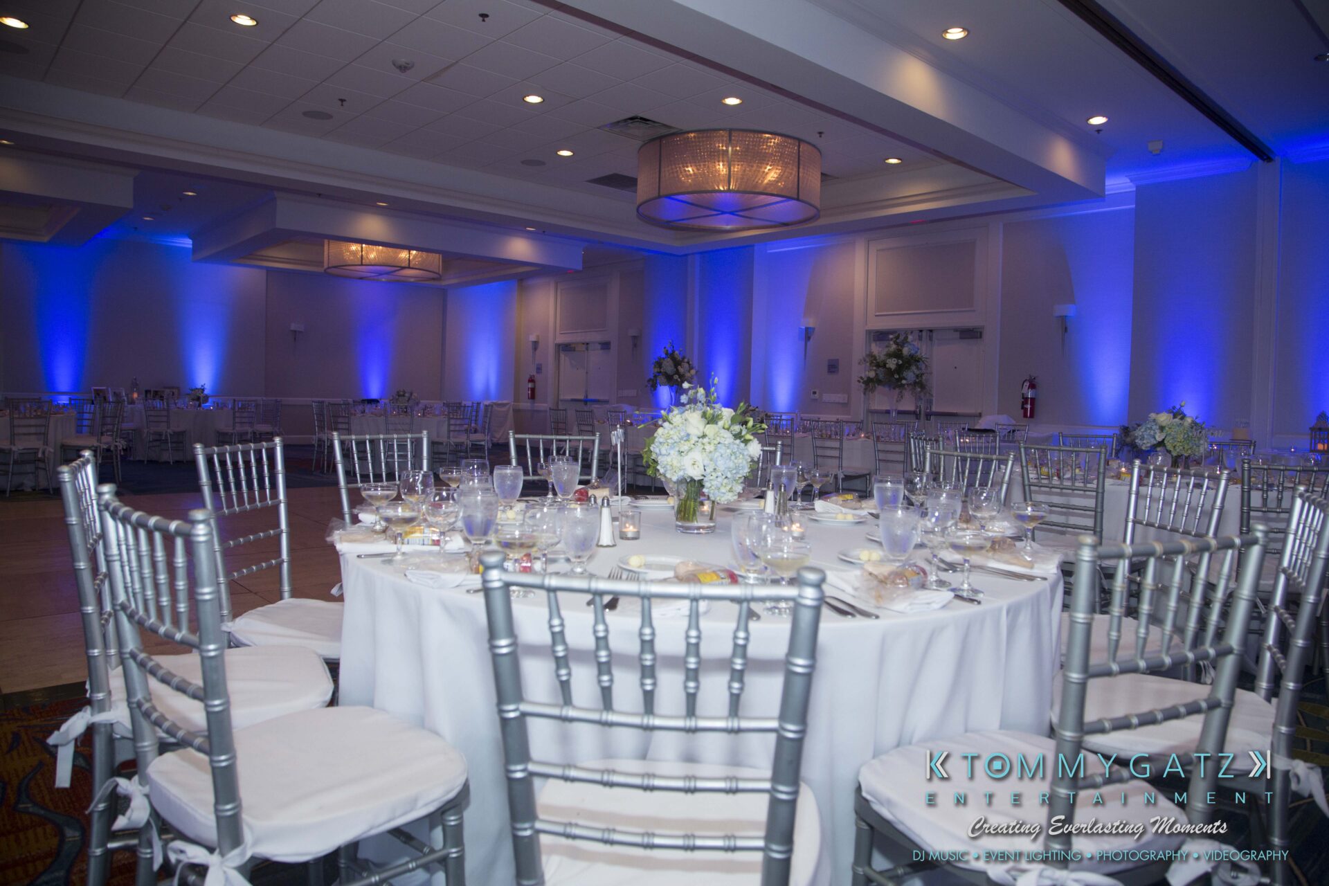 Carly-Patrick-FINAL-Blog-Watermark-13 - TOMMY GATZ ENTERTAINMENT A decorated banquet hall with round tables, floral centerpieces, white table settings, and blue lighting. Silver chairs surround each table.