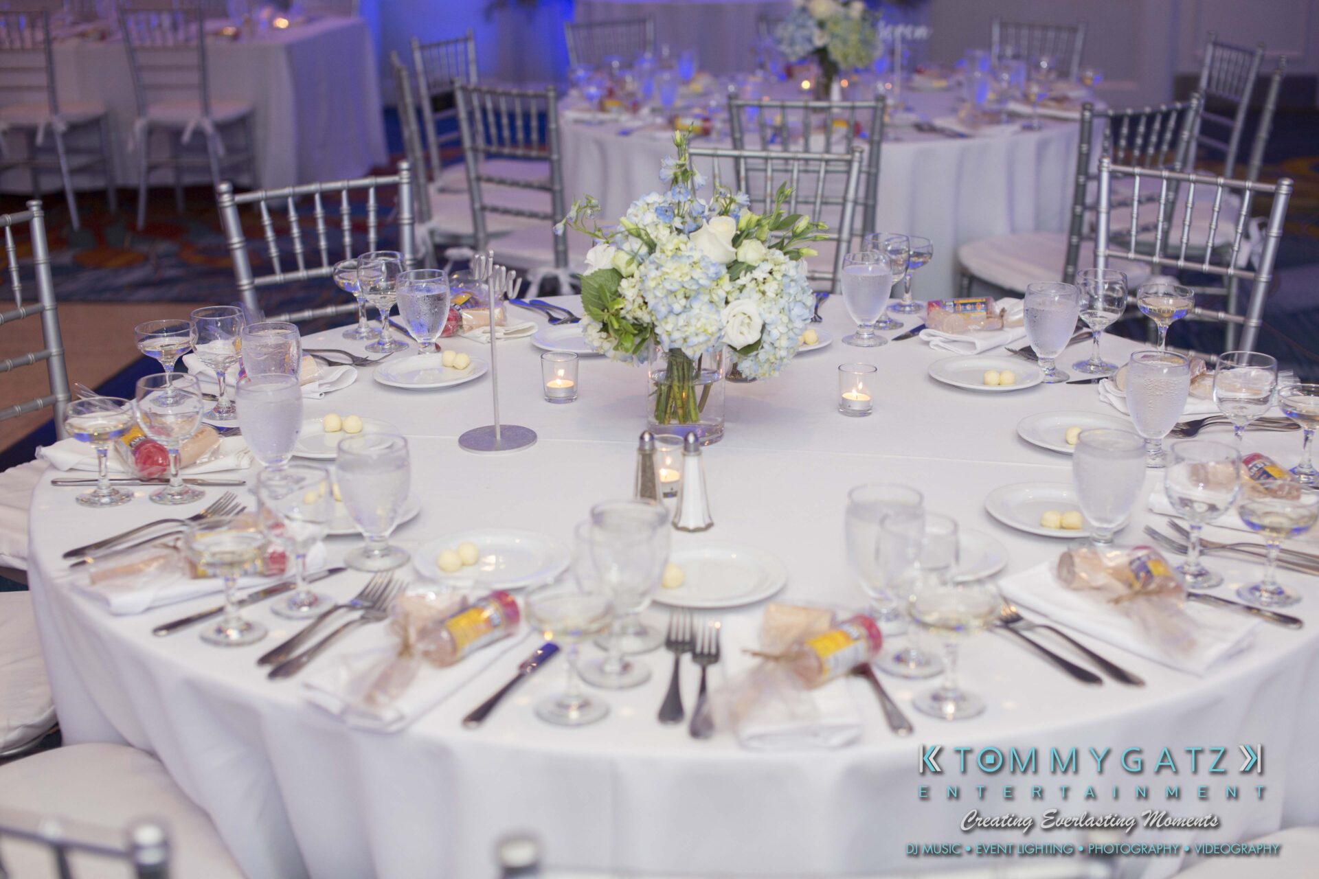 Carly-Patrick-FINAL-Blog-Watermark-21 - TOMMY GATZ ENTERTAINMENT Elegant table setting for a formal event with white tablecloth, floral centerpiece, wine glasses, silverware, small candles, and party favors.