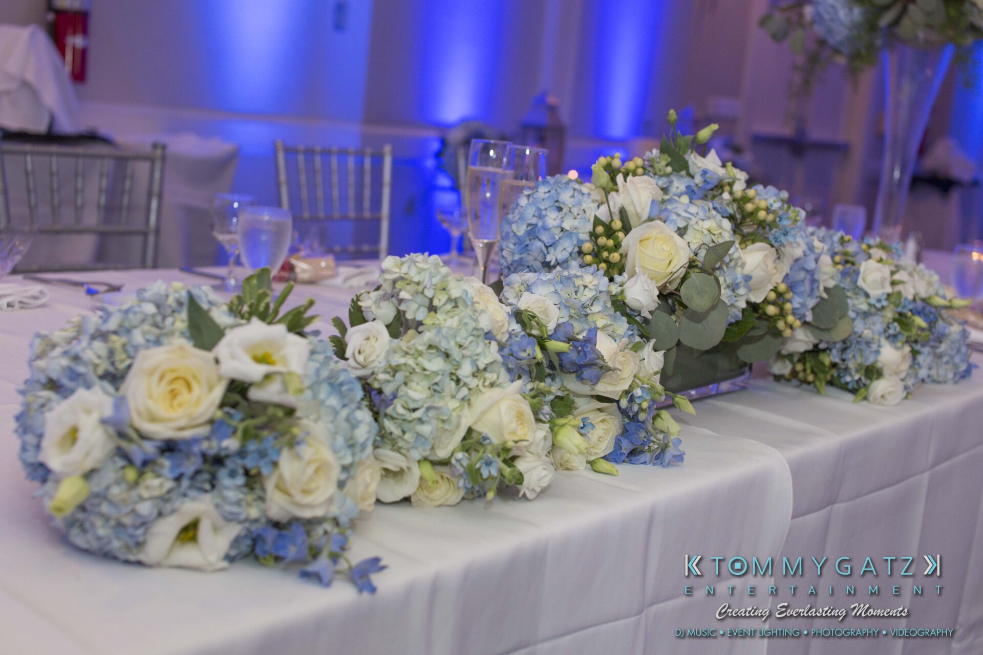 Carly-Patrick-FINAL-Blog-Watermark-24 - TOMMY GATZ ENTERTAINMENT Floral arrangements of blue hydrangeas and white roses on a table with a white tablecloth.