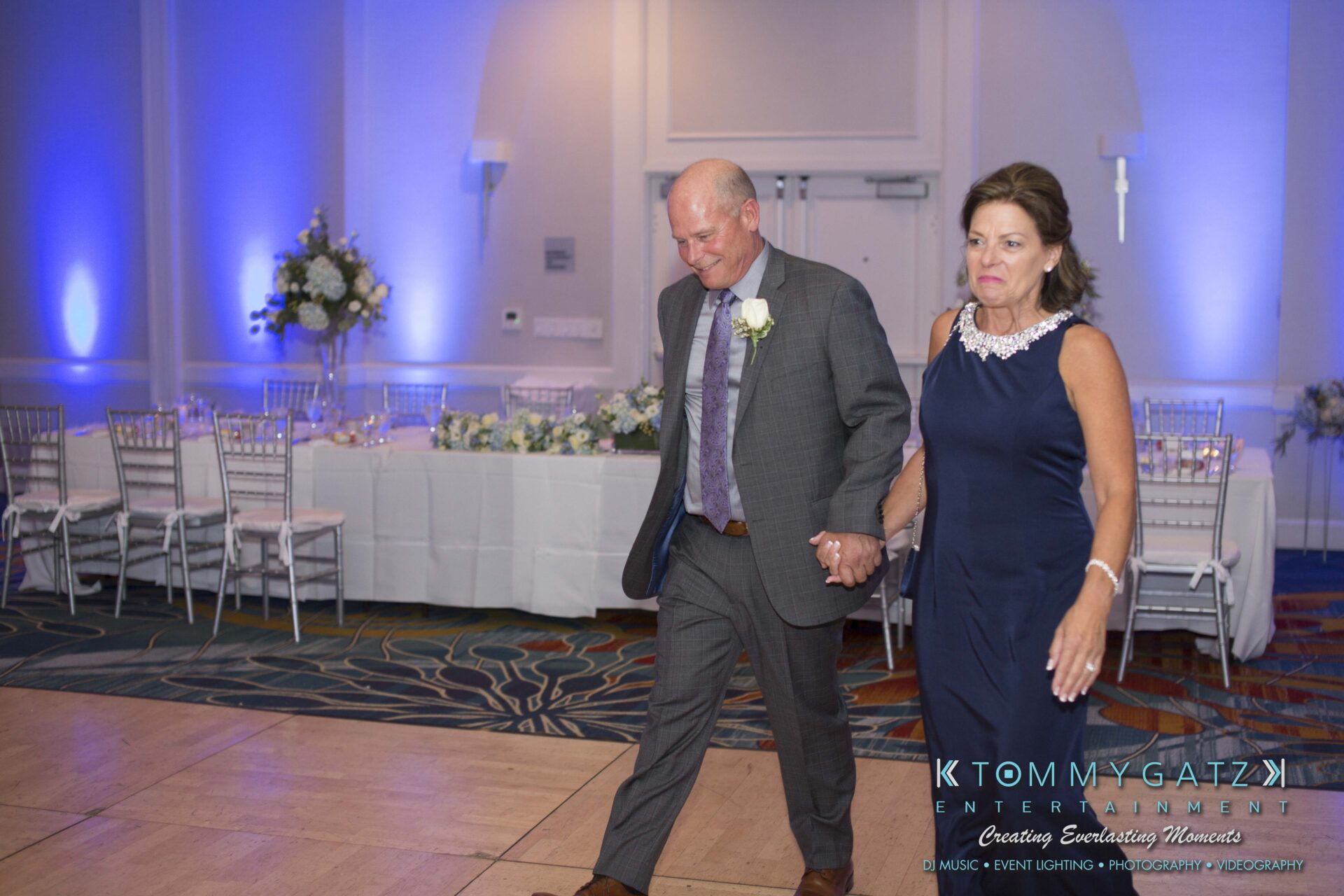 Carly-Patrick-FINAL-Blog-Watermark-26 - TOMMY GATZ ENTERTAINMENT A man and woman holding hands walk across a decorated event space with tables and floral arrangements.