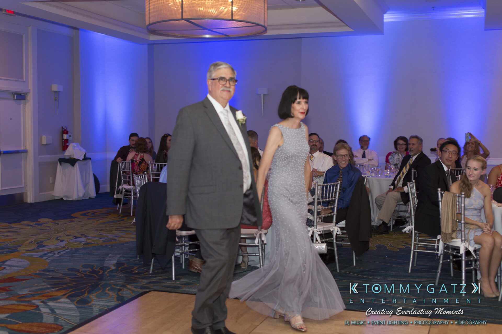 Carly-Patrick-FINAL-Blog-Watermark-27 - TOMMY GATZ ENTERTAINMENT A man in a suit and a woman in a gown walk together in a room with seated guests and blue lighting.