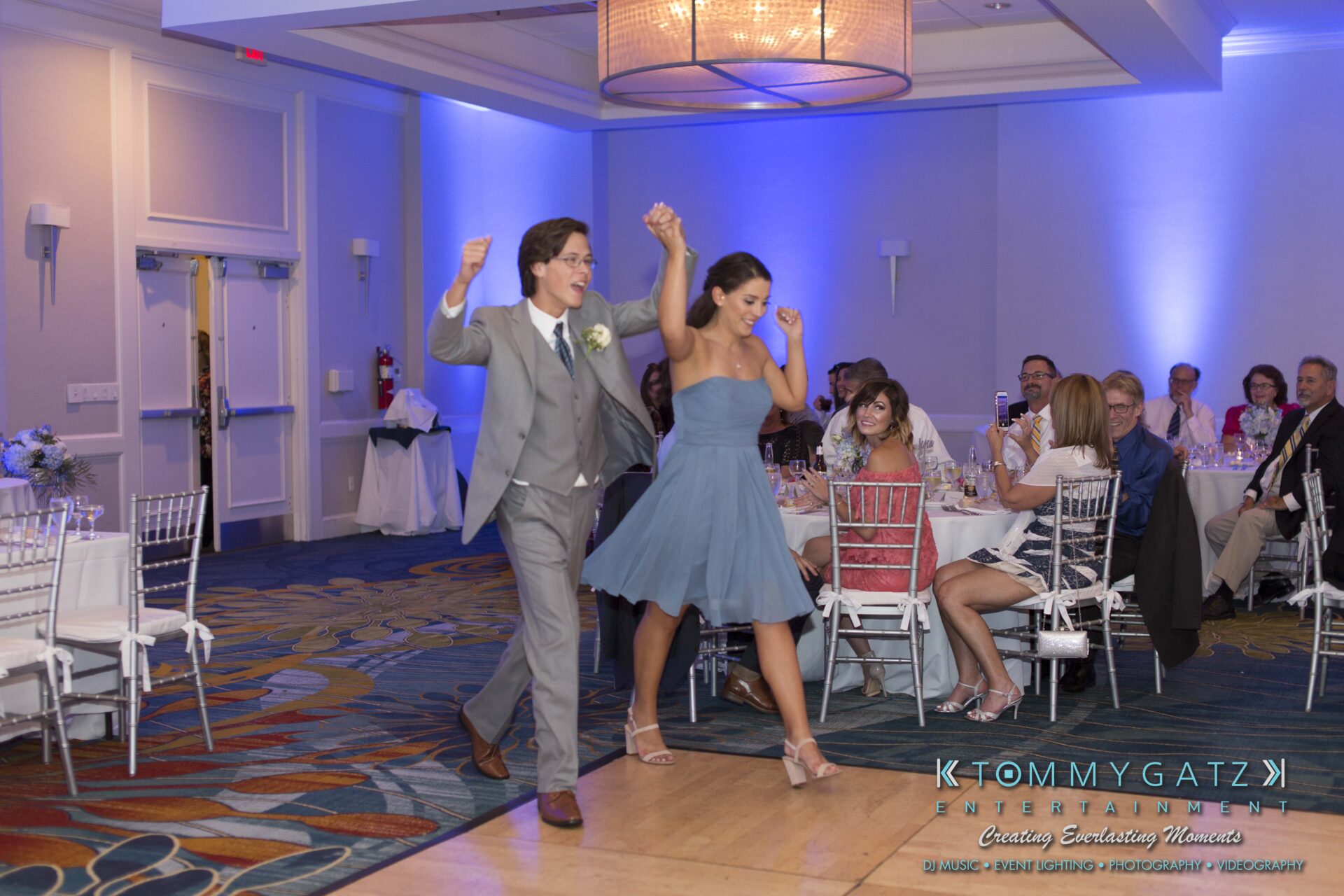 Carly-Patrick-FINAL-Blog-Watermark-30 - TOMMY GATZ ENTERTAINMENT A man in a gray suit and sunglasses and a woman in a blue dress and heels dance into a wedding reception hall, with seated guests watching.