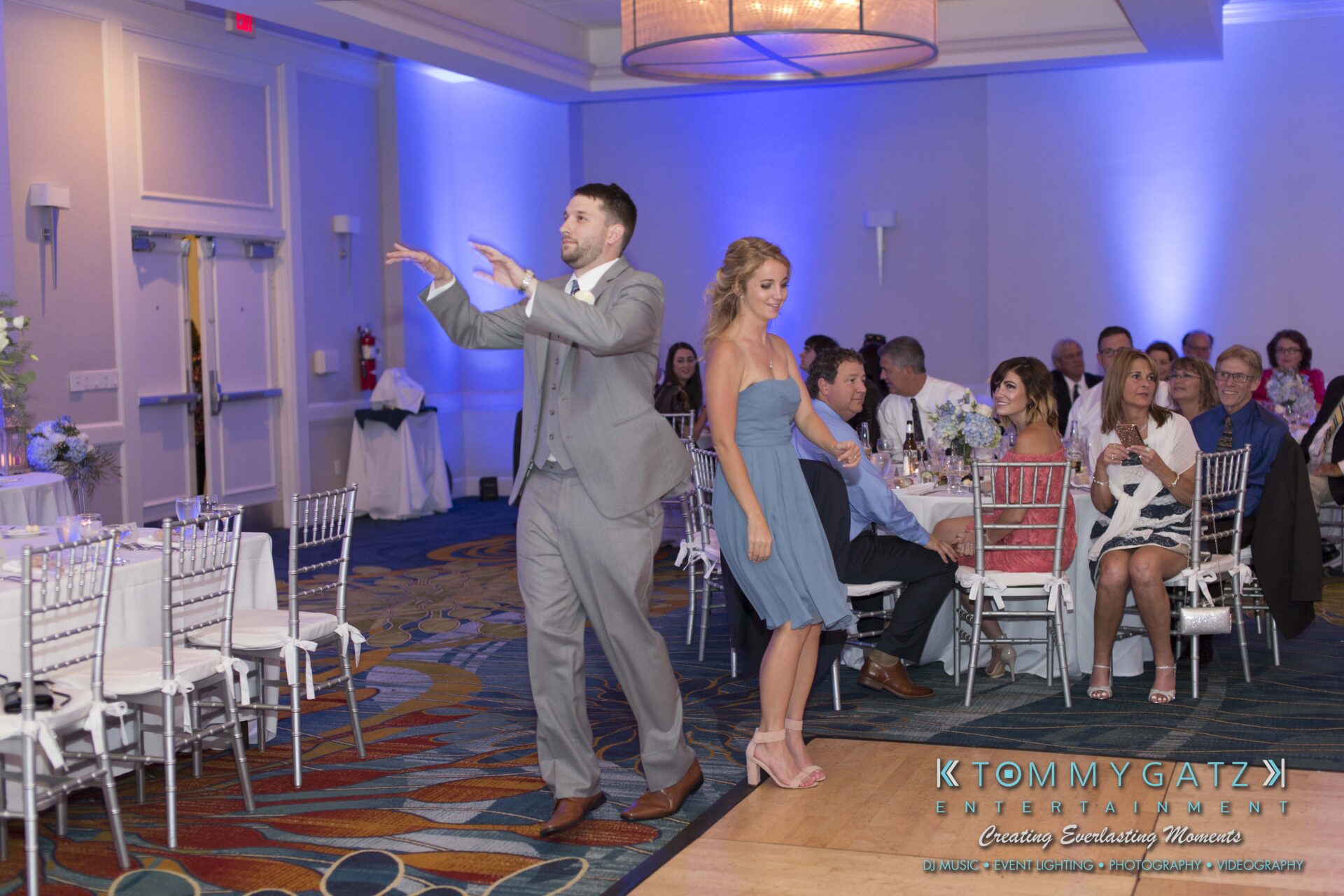 Carly-Patrick-FINAL-Blog-Watermark-31 - TOMMY GATZ ENTERTAINMENT A man and woman in formal attire are walking and posing playfully in a decorated banquet hall with seated guests watching.