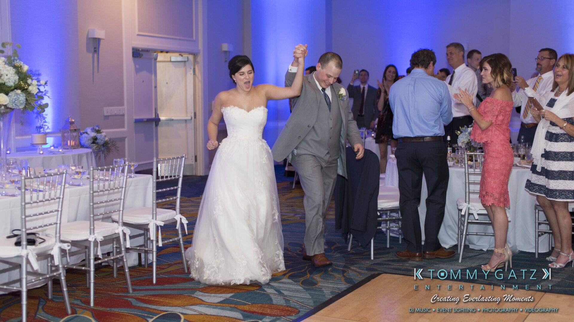 Carly-Patrick-FINAL-Blog-Watermark-33 - TOMMY GATZ ENTERTAINMENT A bride and groom celebrate while entering a decorated reception hall, with guests clapping.