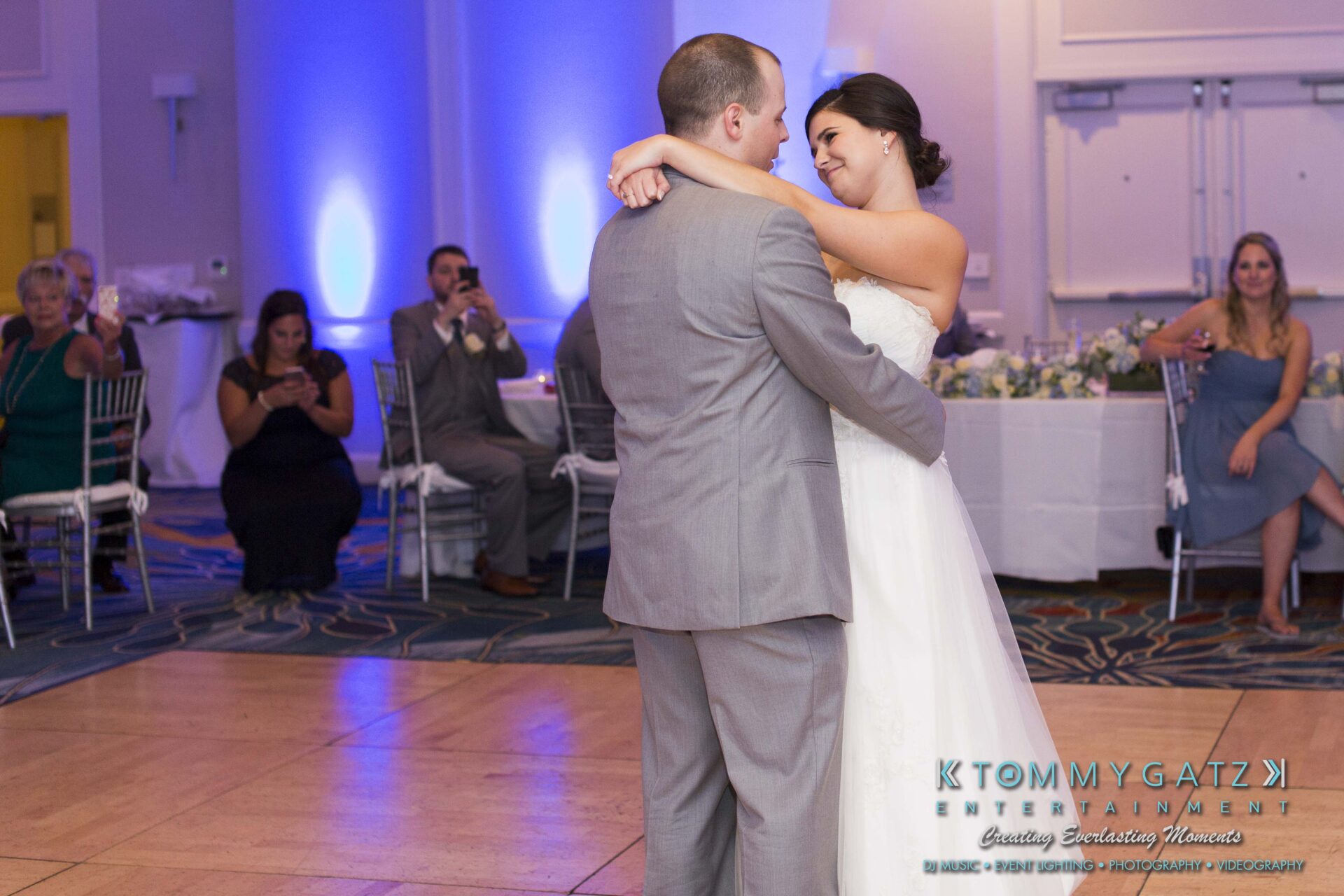 Carly-Patrick-FINAL-Blog-Watermark-34 - TOMMY GATZ ENTERTAINMENT A bride and groom share a first dance at a wedding reception. Guests are seated around the dance floor, with one taking photos. Blue lighting adds ambiance to the room.