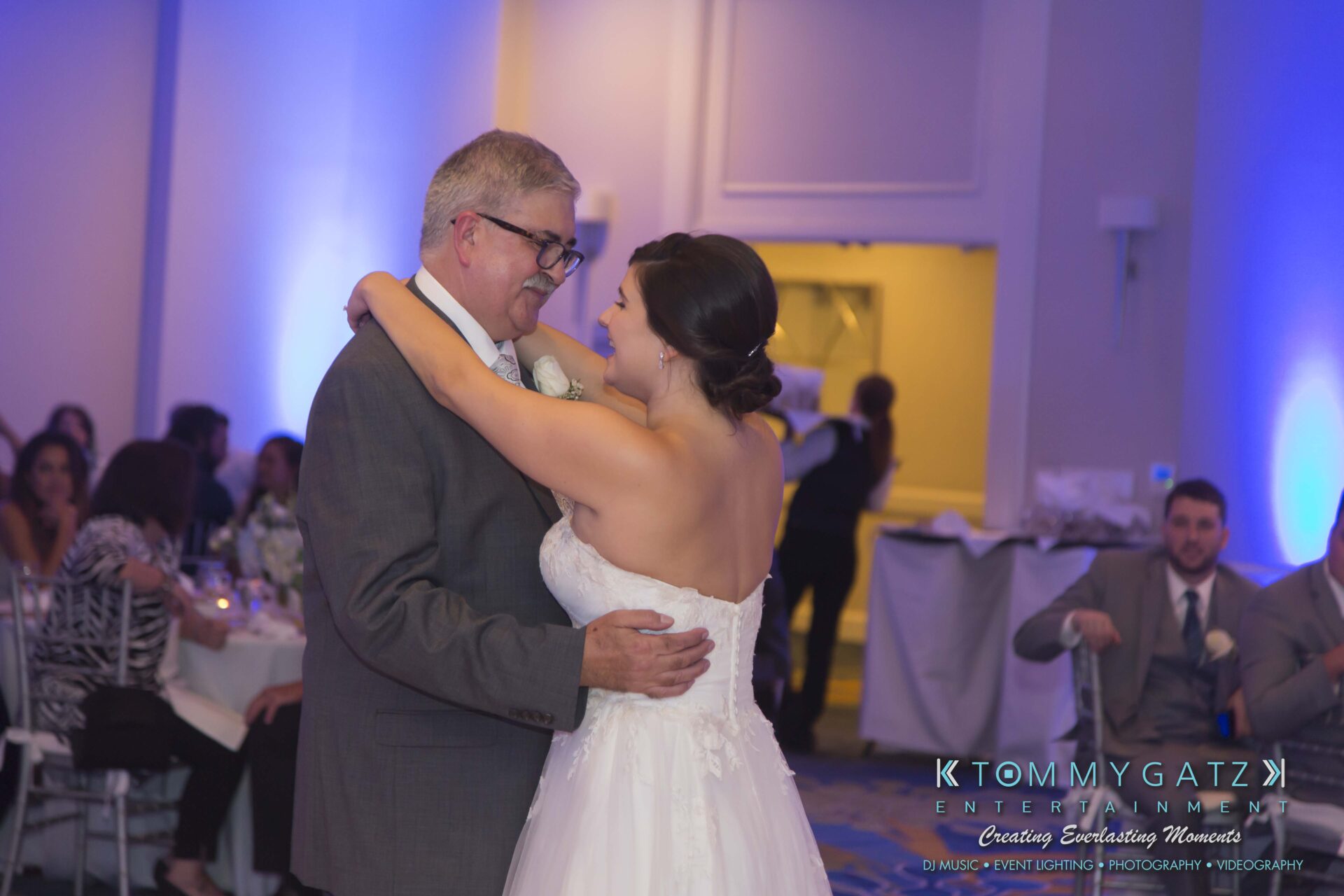 Carly-Patrick-FINAL-Blog-Watermark-36 - TOMMY GATZ ENTERTAINMENT A bride and an older man smile while dancing together in a banquet hall. Guests sit at tables in the background.