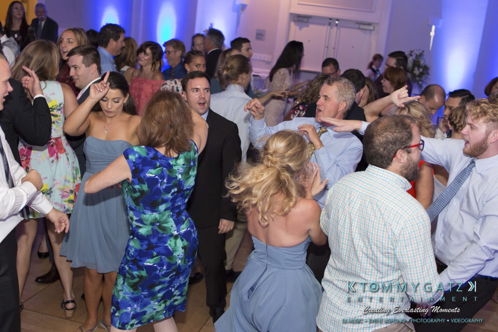 Carly-Patrick-FINAL-Blog-Watermark-38 - TOMMY GATZ ENTERTAINMENT A group of people in formal and semi-formal attire are dancing energetically in a spacious indoor venue with blue ambient lighting.