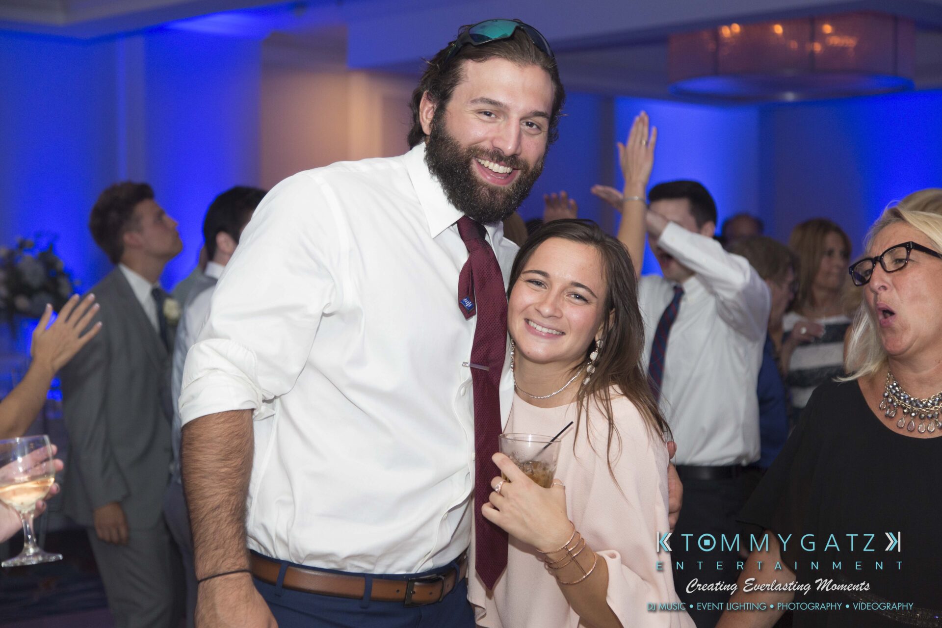 Carly-Patrick-FINAL-Blog-Watermark-40 - TOMMY GATZ ENTERTAINMENT A man and woman pose together at a party, smiling. People are dancing in the background.