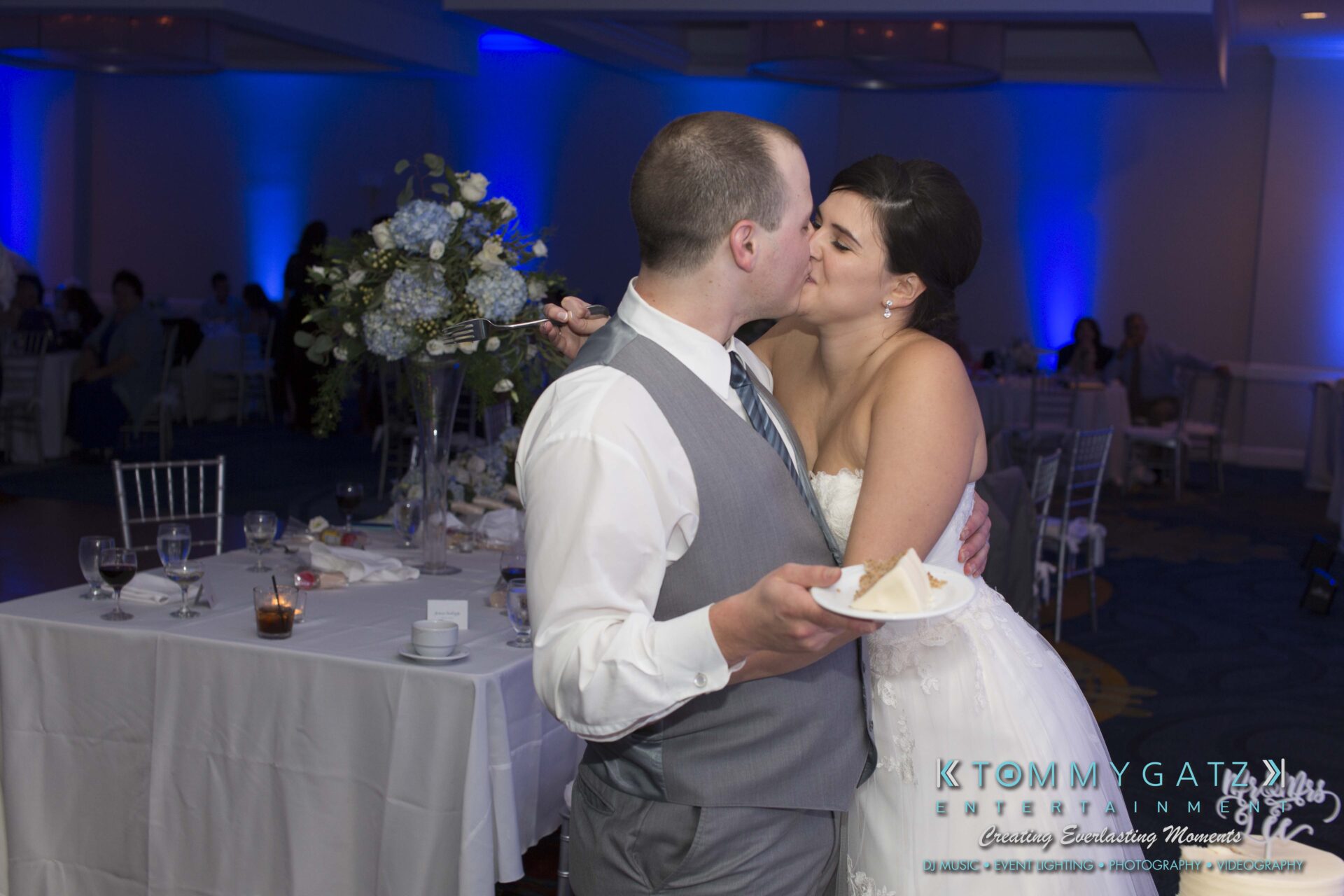 Carly-Patrick-FINAL-Blog-Watermark-44 - TOMMY GATZ ENTERTAINMENT Bride and groom share a kiss while holding cake slices in a decorated reception hall.