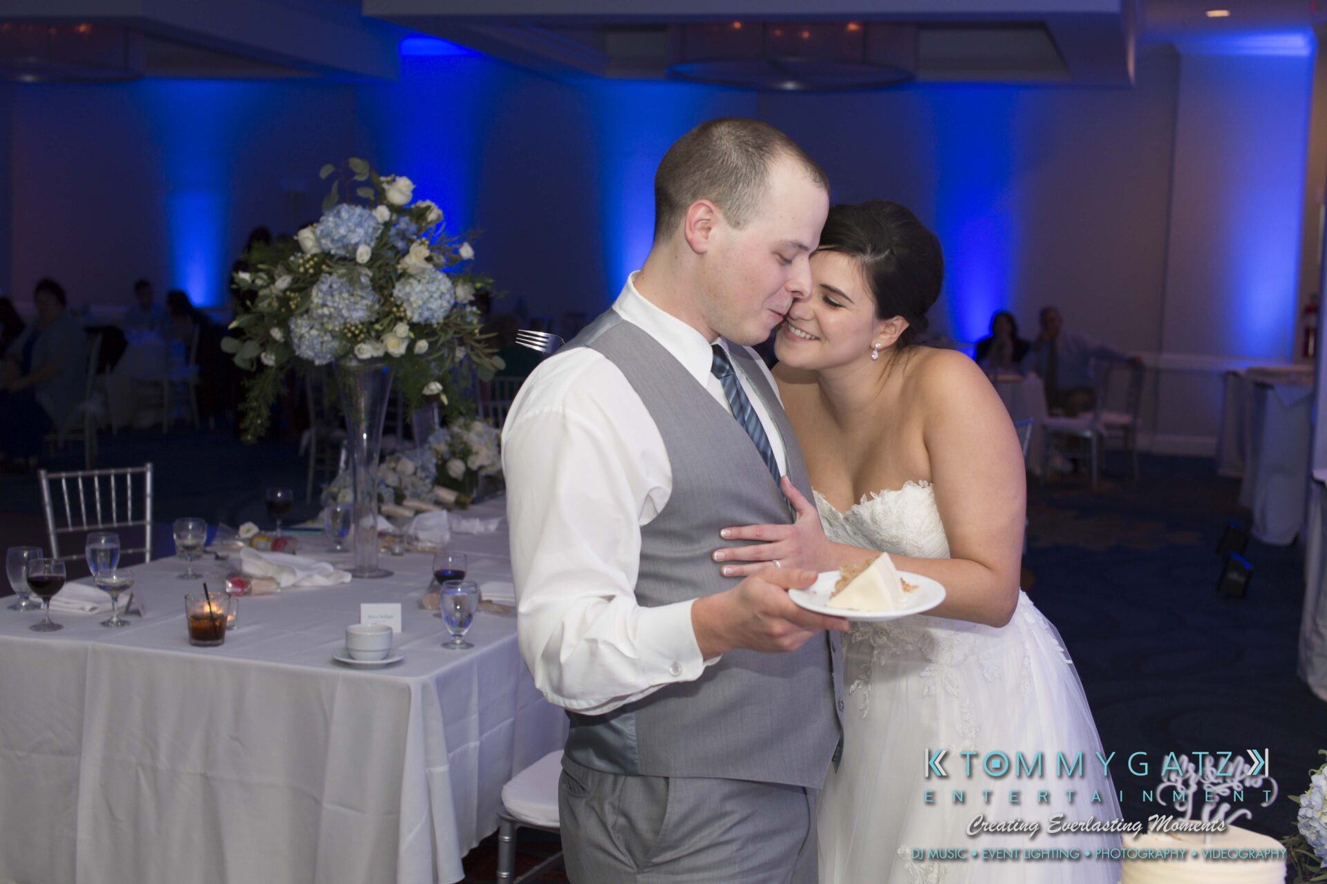 Carly-Patrick-FINAL-Blog-Watermark-45 - TOMMY GATZ ENTERTAINMENT A couple embraces near a table set with candles and flowers at a wedding reception. The groom holds a plate with a slice of cake.