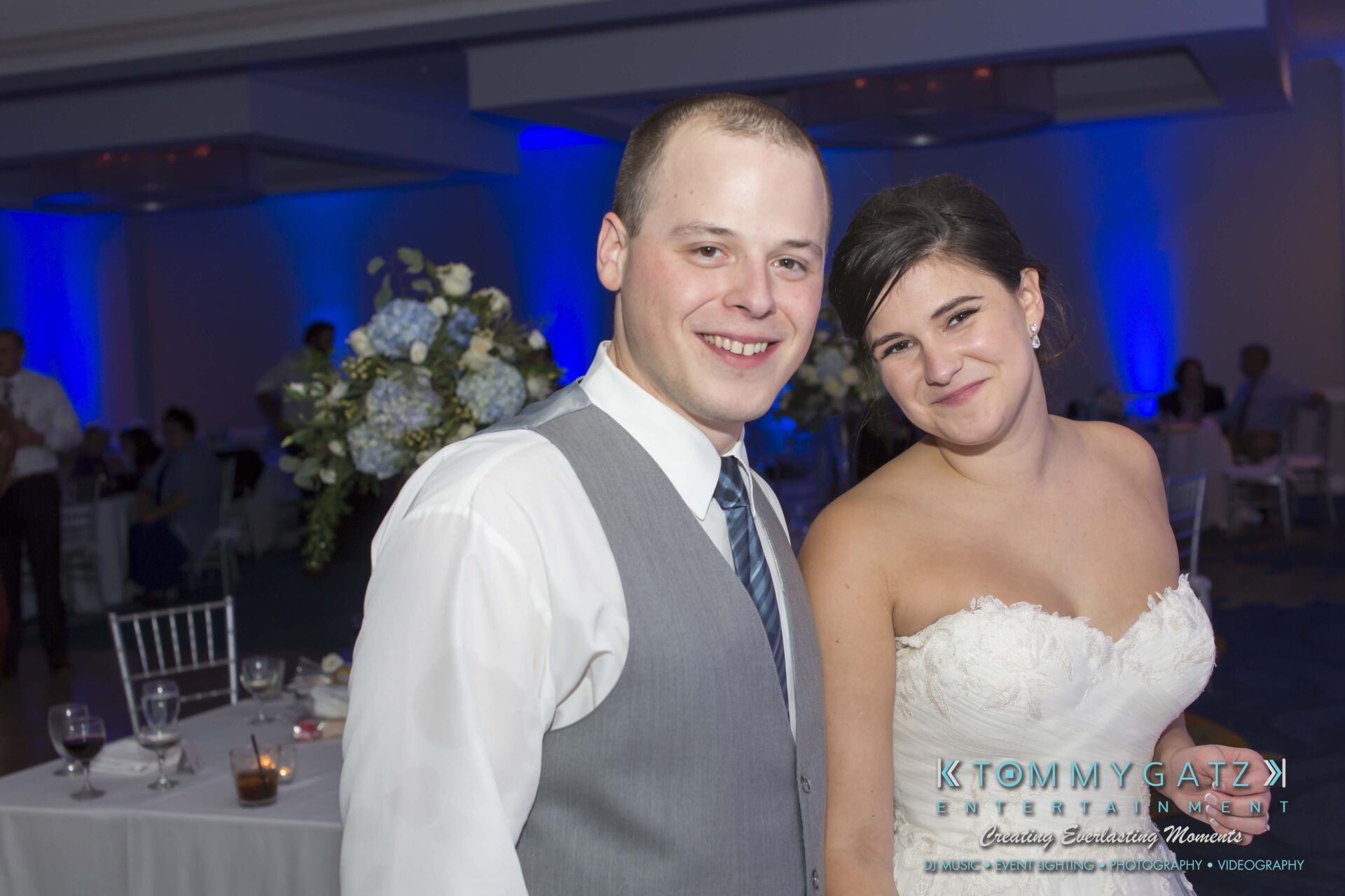 Carly-Patrick-FINAL-Blog-Watermark-46 - TOMMY GATZ ENTERTAINMENT A couple smiling at a formal event, with the woman in a white strapless dress and the man in a gray vest and tie. The setting features dim lighting and floral decor.