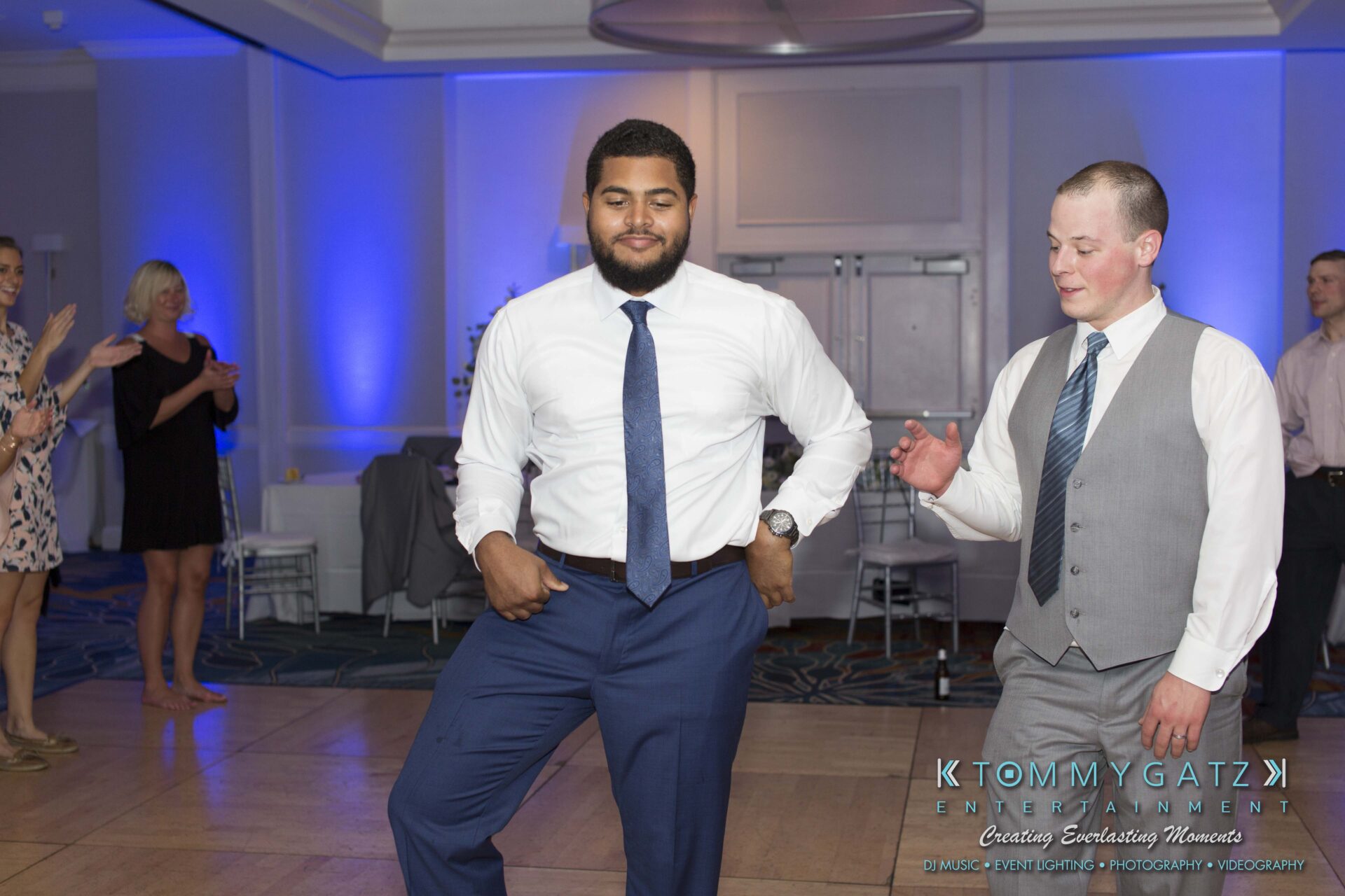 Carly-Patrick-FINAL-Blog-Watermark-49 - TOMMY GATZ ENTERTAINMENT Two men are dancing on a wooden floor at a party, wearing formal attire. Others watch and clap in the background.