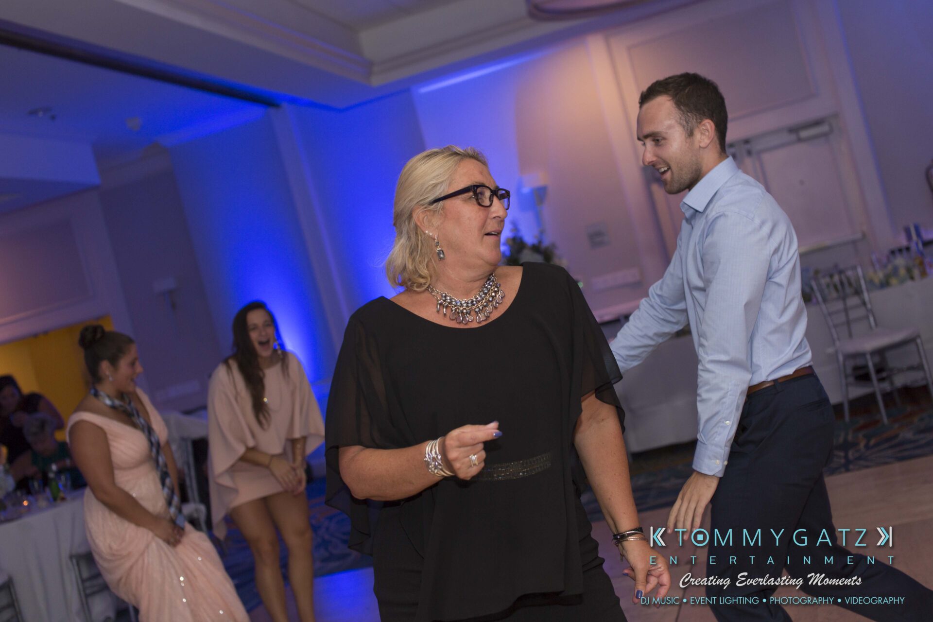 Carly-Patrick-FINAL-Blog-Watermark-50 - TOMMY GATZ ENTERTAINMENT People dancing and smiling in a dimly lit room with blue lighting.